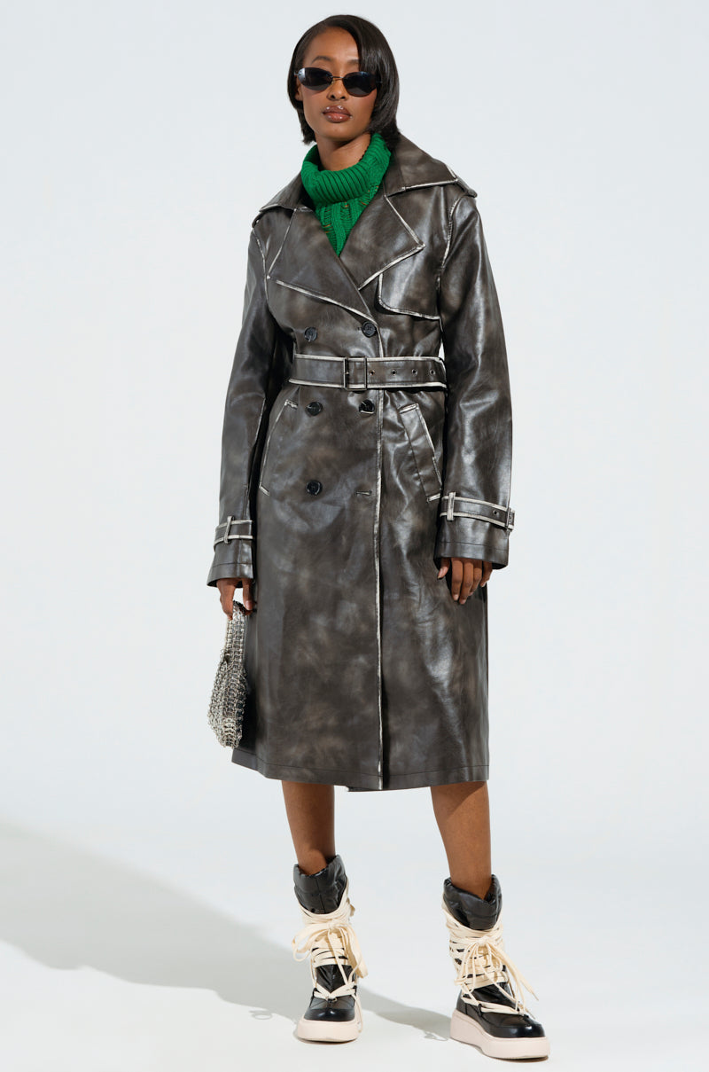 A person stands confidently in an ACID RAP FAUX LEATHER TRENCH, characterized by its dark, glossy finish. Underneath, they wear a green turtleneck sweater and accessorize with sunglasses. They hold a small handbag and complete the look with stylish high-top boots. The plain background emphasizes the trench coat's double-breasted button closures, highlighting the fashion ensemble.