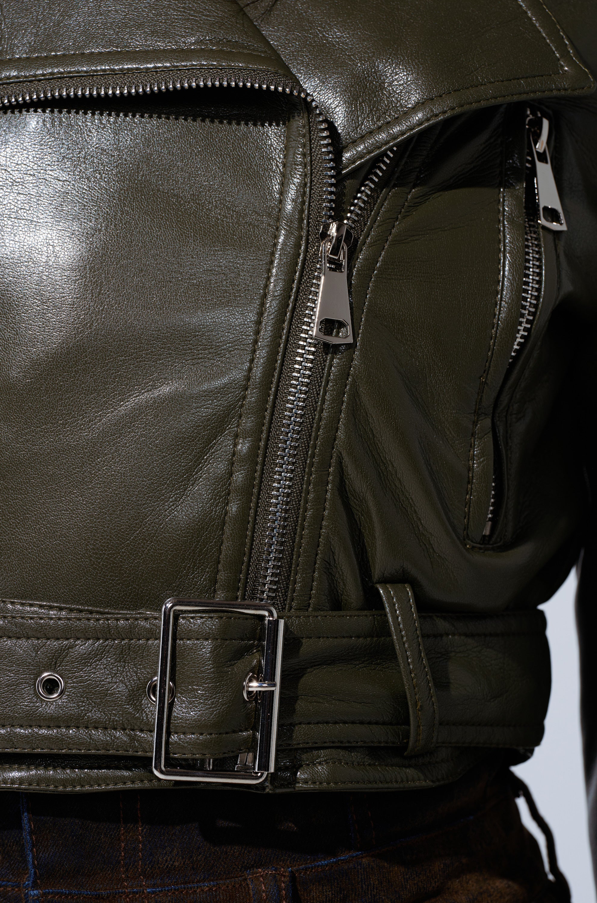 Close-up of the BUTTER LUXE EVERYDAY MOTO jacket in olive green, showcasing its shiny silver zipper, matching buckle belt, zippered pocket, and luxe faux leather texture with crisp metal details.