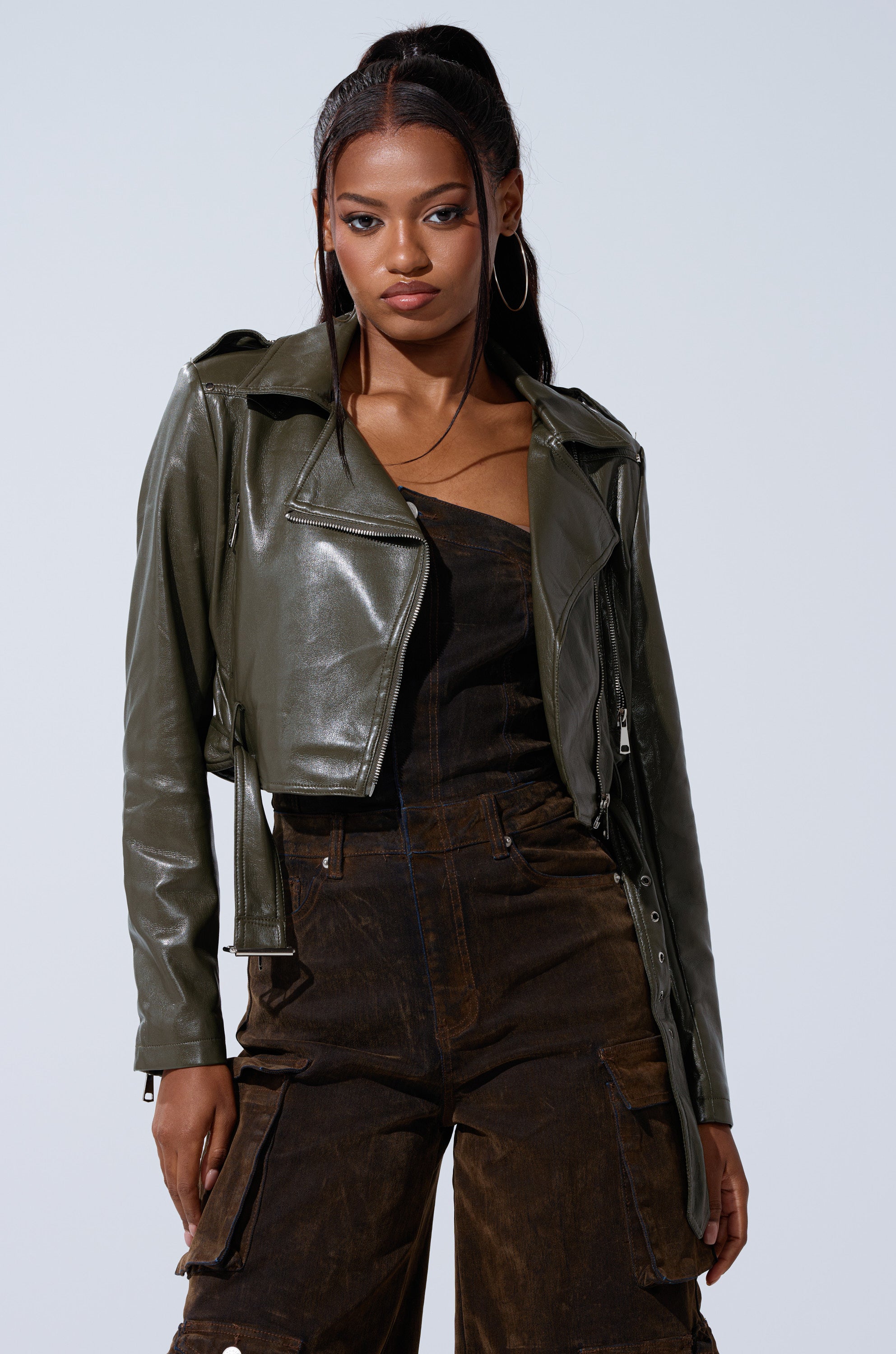 A woman with long dark hair in a ponytail and hoop earrings wears the shiny dark green BUTTER LUXE EVERYDAY MOTO cropped jacket with an adjustable buckle, over a brown one-shoulder jumpsuit, standing against a plain light background.