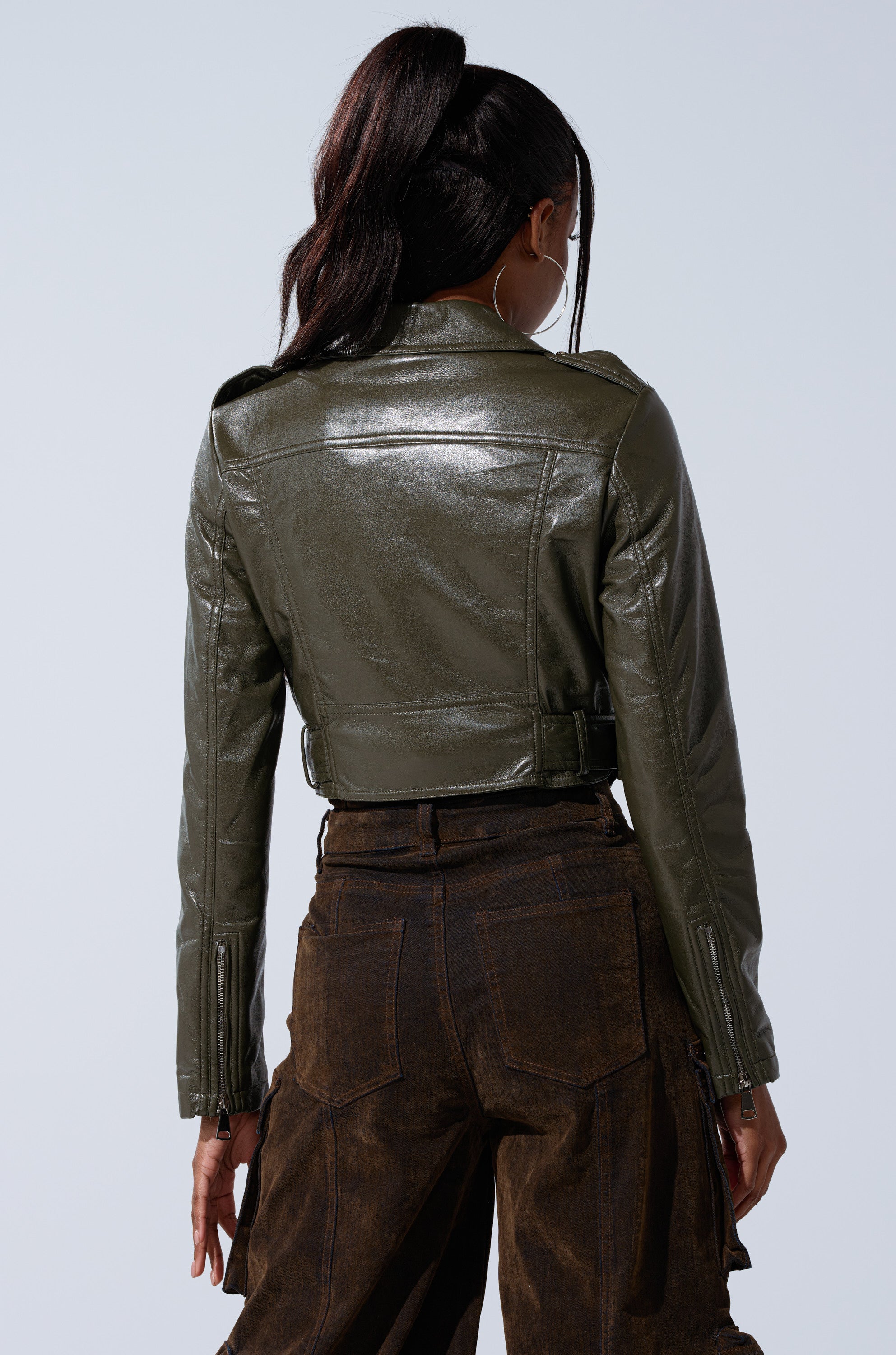 A person with long dark hair in a ponytail wears the BUTTER LUXE EVERYDAY MOTO jacket in olive green and brown high-waisted pants, facing away from the camera against a light gray background.