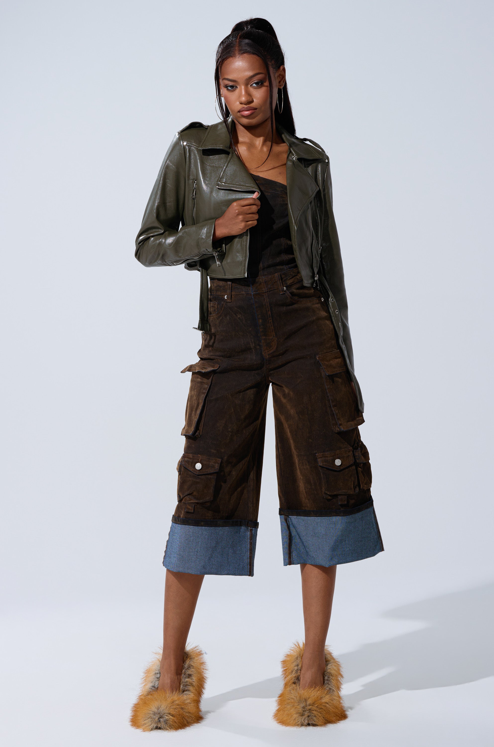 A woman poses confidently in the BUTTER LUXE EVERYDAY MOTO faux leather jacket with an adjustable buckle belt, styled with a black top, brown cargo culotte pants featuring denim cuffs, and fuzzy orange slippers against a plain light background.