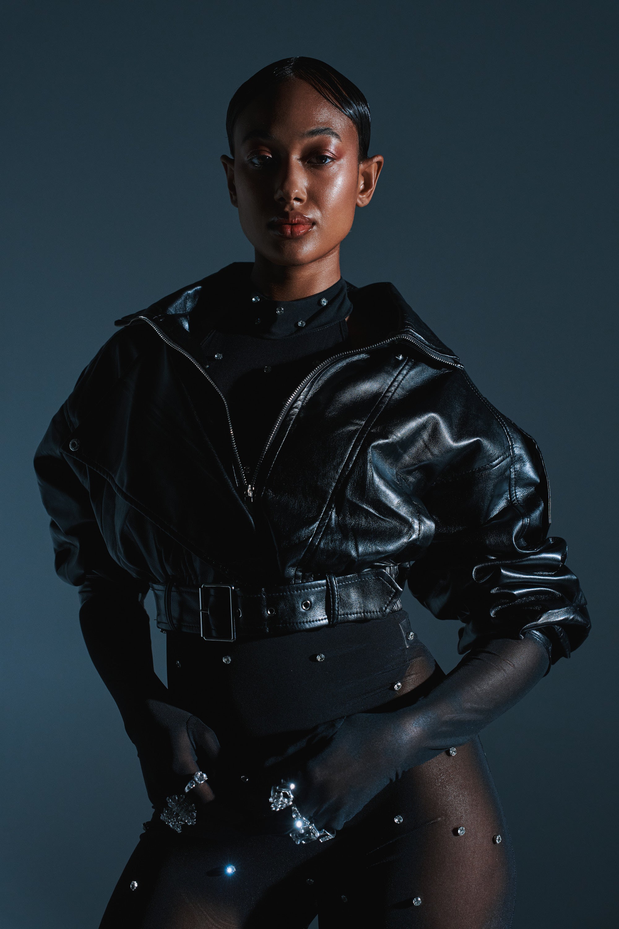 A person poses confidently in the EVA SLOUCHY MOTO IN BLACK jacket, styled with sheer embellished pants and statement rings, set against a dark blue background with dramatic lighting. 2