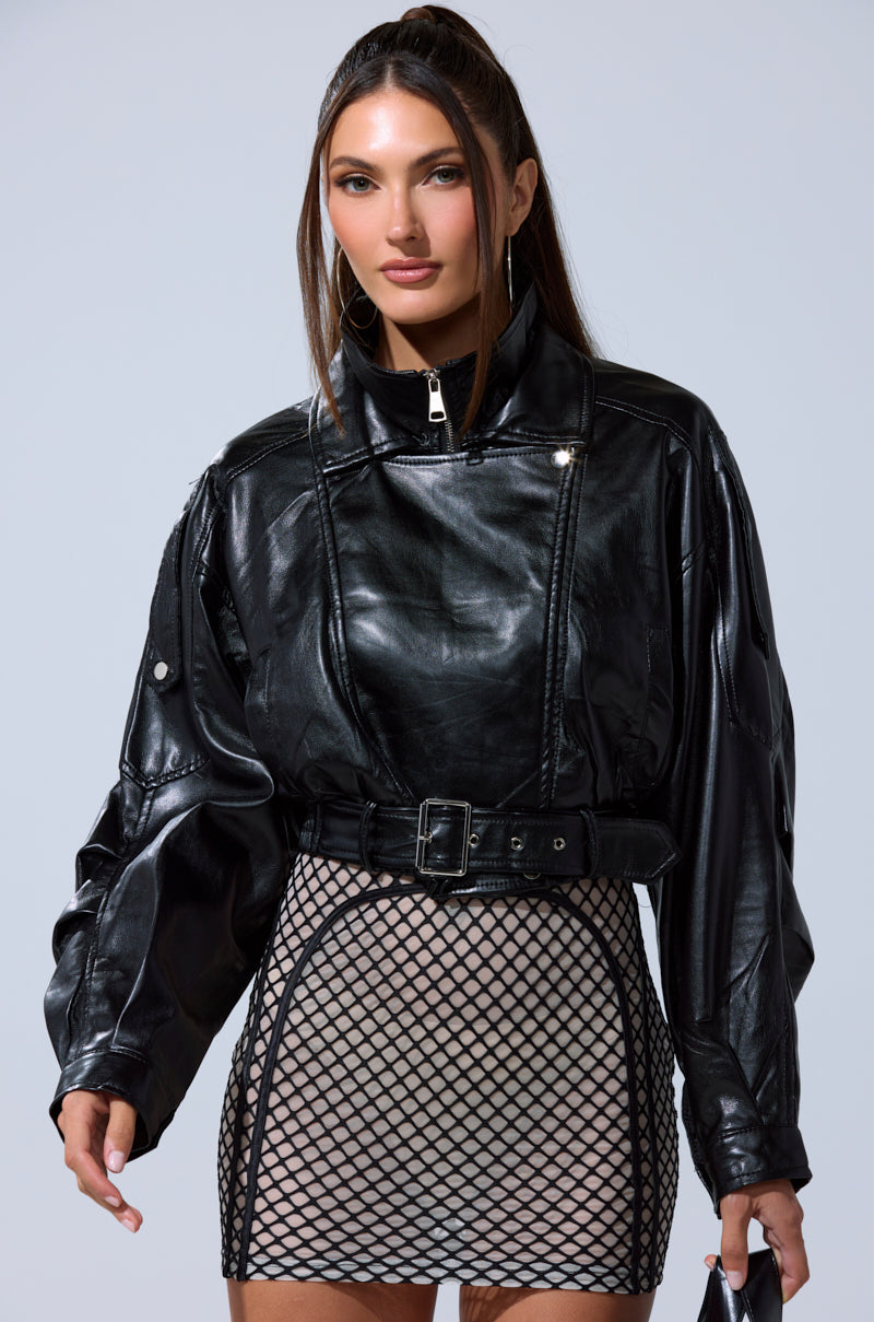 A woman models the EVA SLOUCHY MOTO IN BLACK, a cropped oversized leather jacket, styled over a beige mini skirt with a black fishnet overlay against a plain light background. 6