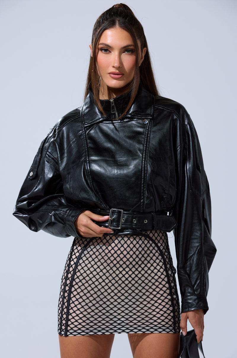 A woman wearing the EVA SLOUCHY MOTO IN BLACK vegan leather jacket with a belt and a beige mini skirt over black fishnet mesh stands against a light gray background. 7