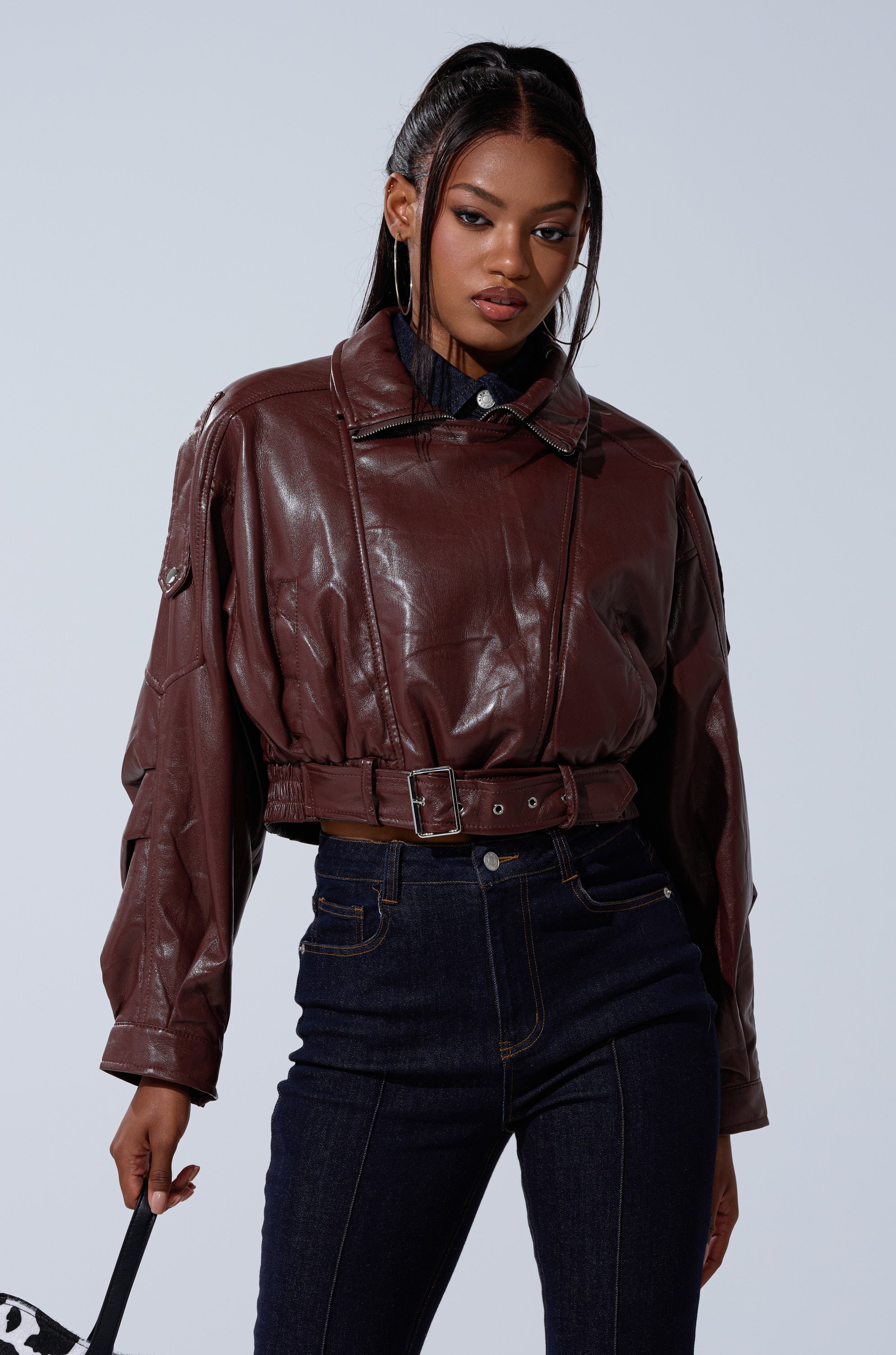 A woman poses confidently against a plain light background, wearing the shiny brown EVA SLOUCHY MOTO faux leather jacket with high-waisted dark jeans, holding a bag. Her long, sleek braids complete the bold look. 1