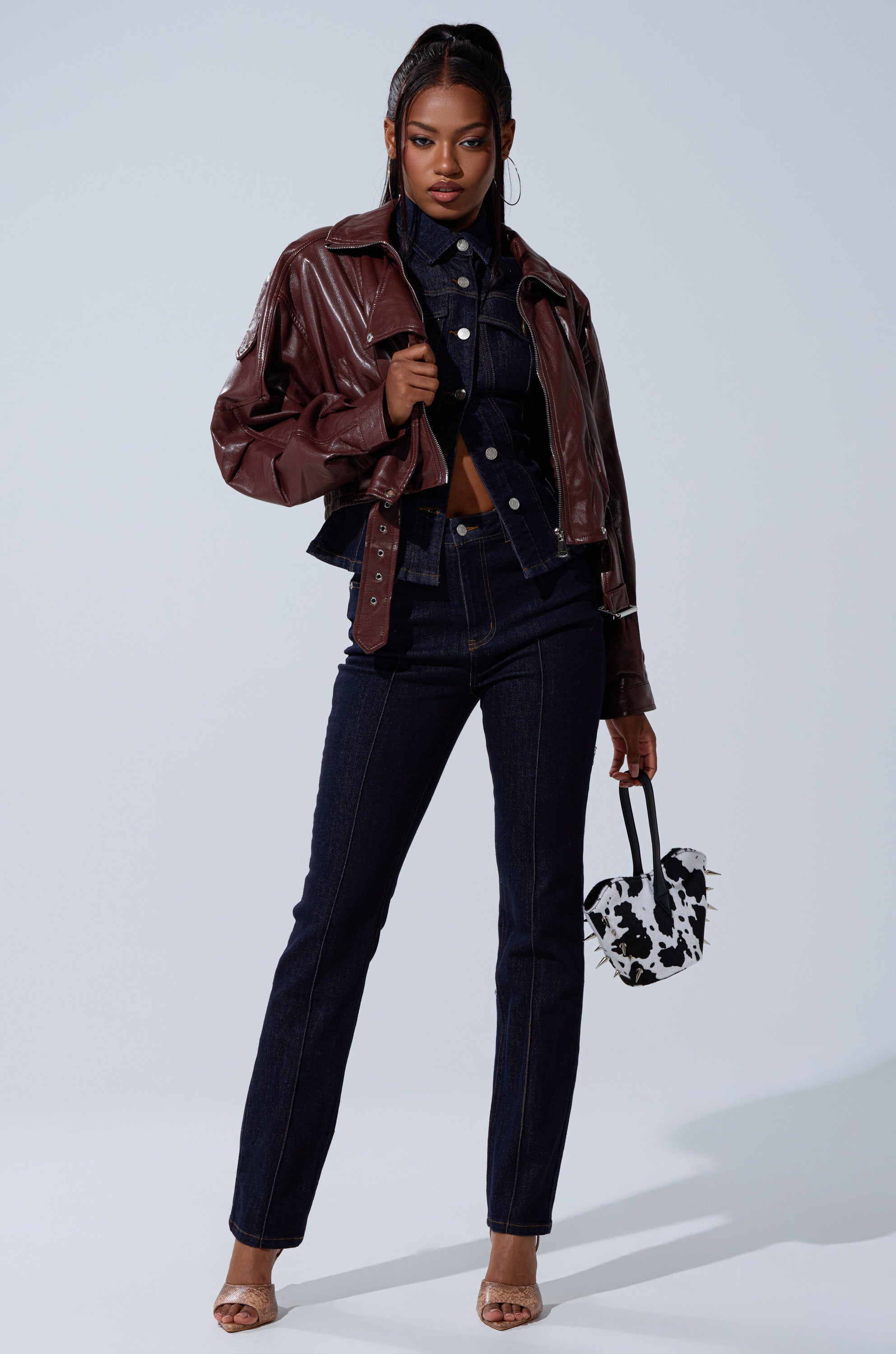 A woman stands confidently in the EVA SLOUCHY MOTO jacket over dark denim straight-leg jeans and a cropped top, holding a black-and-white cow-print handbag and wearing open-toe heels. 10
