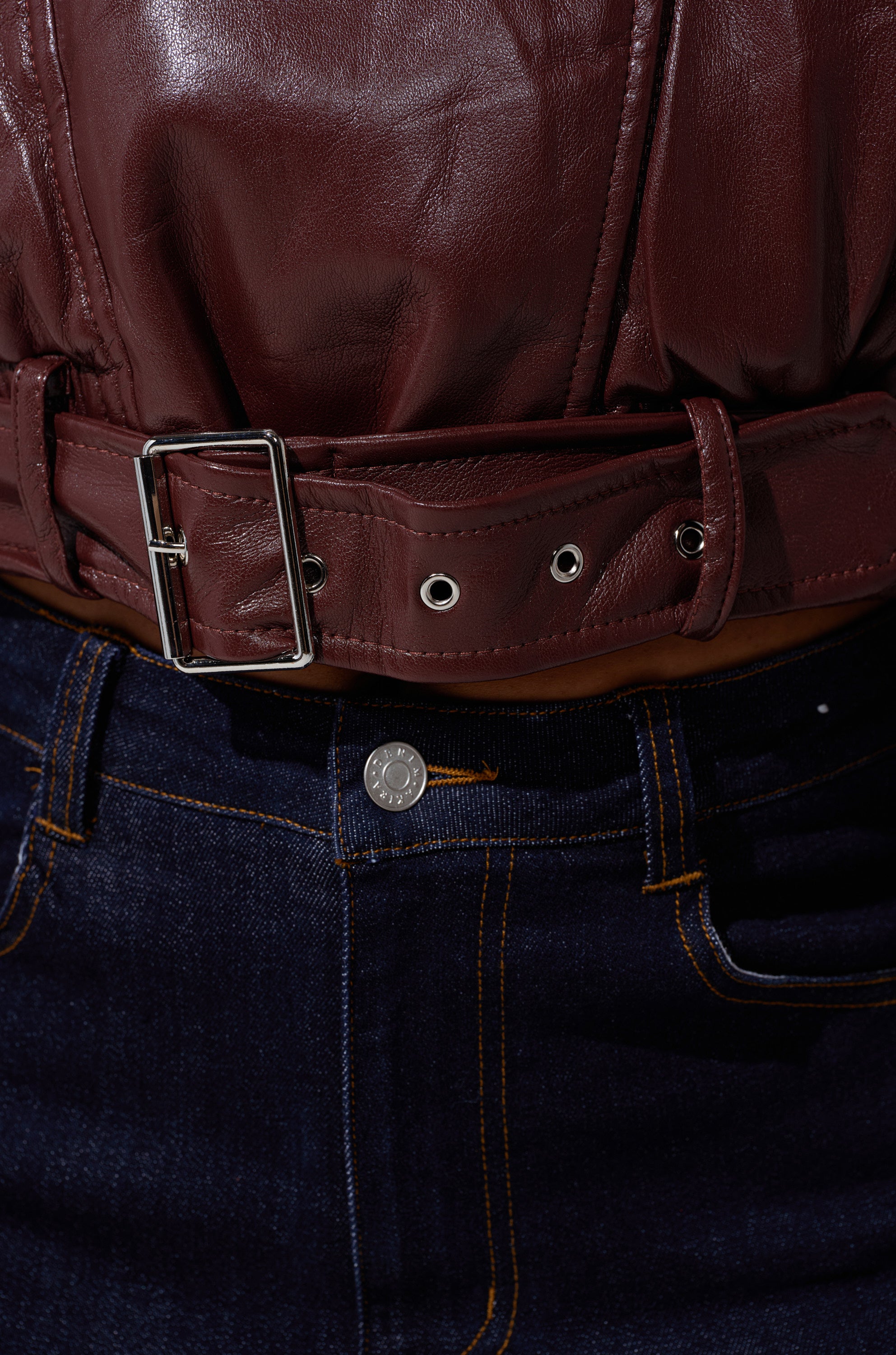 Close-up of someone wearing dark blue denim jeans with orange stitching and a shiny metal button, paired with the EVA SLOUCHY MOTO maroon faux leather jacket featuring a belt and silver buckle. 11