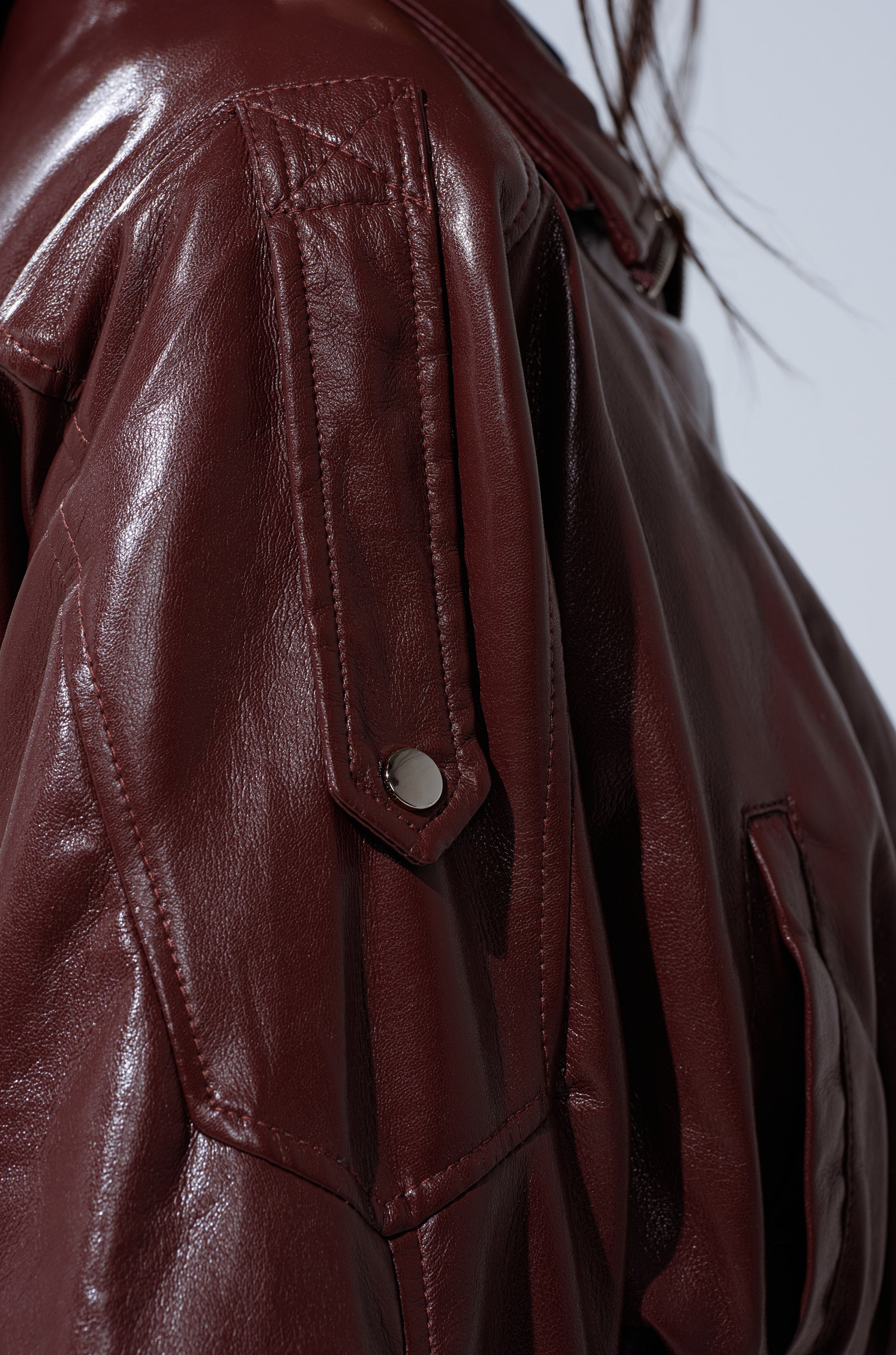 Close-up of the EVA SLOUCHY MOTO jacket in shiny dark brown faux leather, highlighting the shoulder, sleeve seam, snap-button flap pocket, and adjustable belt. Part of a person’s dark hair appears on the right. 12