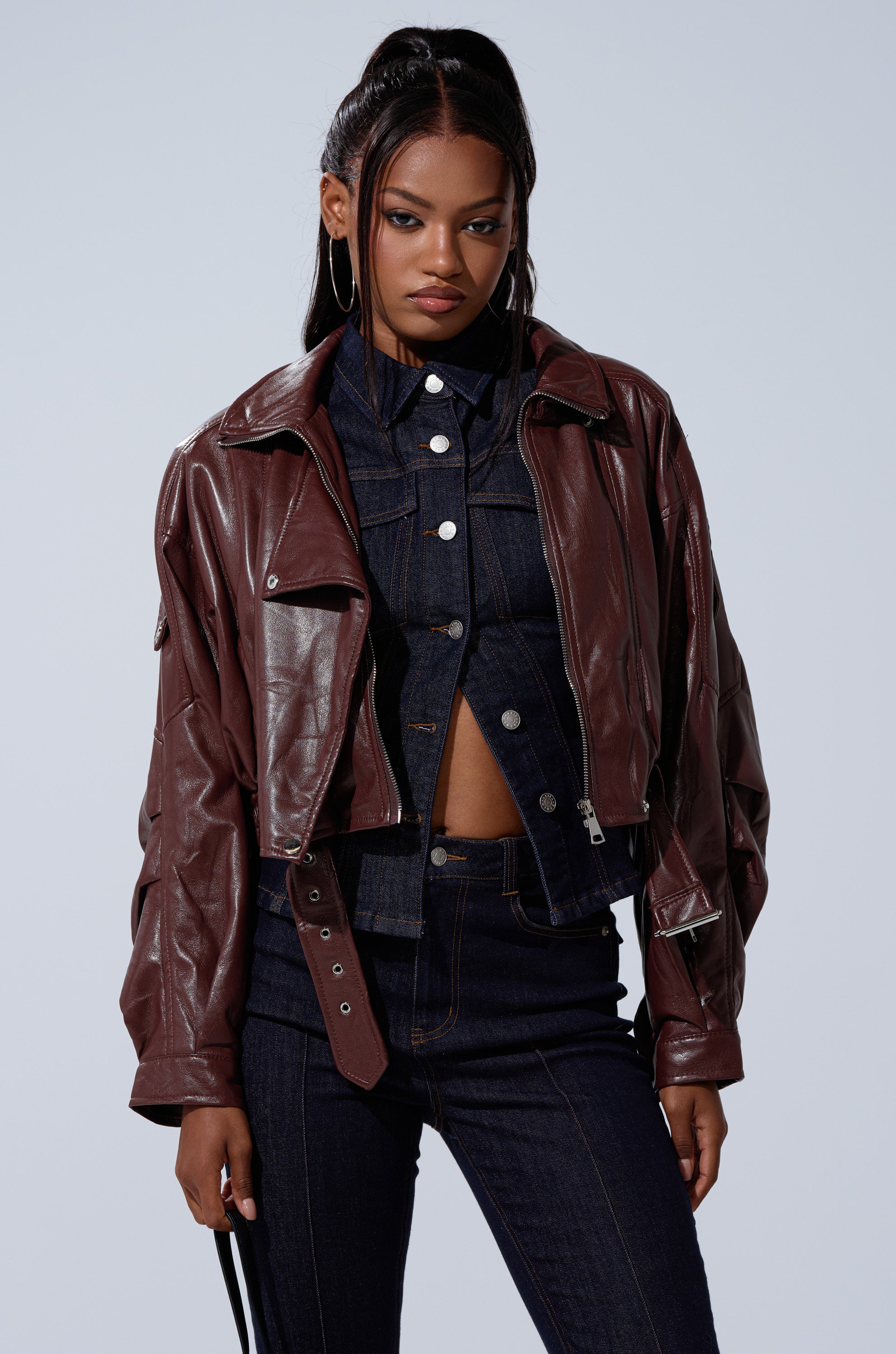 A woman with long, dark hair in a high ponytail wears the EVA SLOUCHY MOTO jacket over a buttoned denim shirt and high-waisted jeans. She stands against a plain light background, confidently looking at the camera. 2