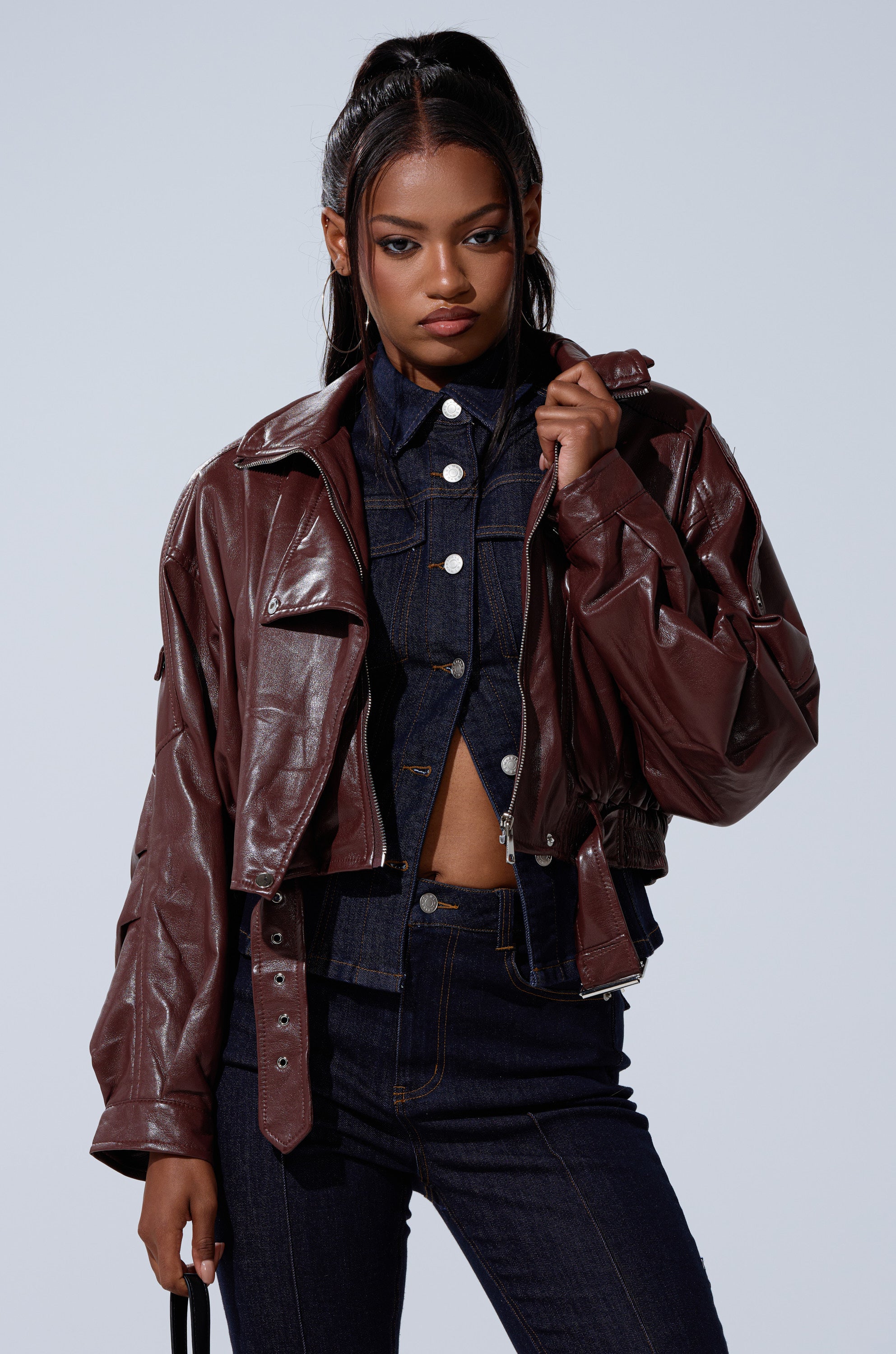 A woman stands against a plain background wearing the EVA SLOUCHY MOTO faux leather jacket over a dark denim button-up shirt and jeans, holding a strap with one hand and gazing confidently at the camera. 3
