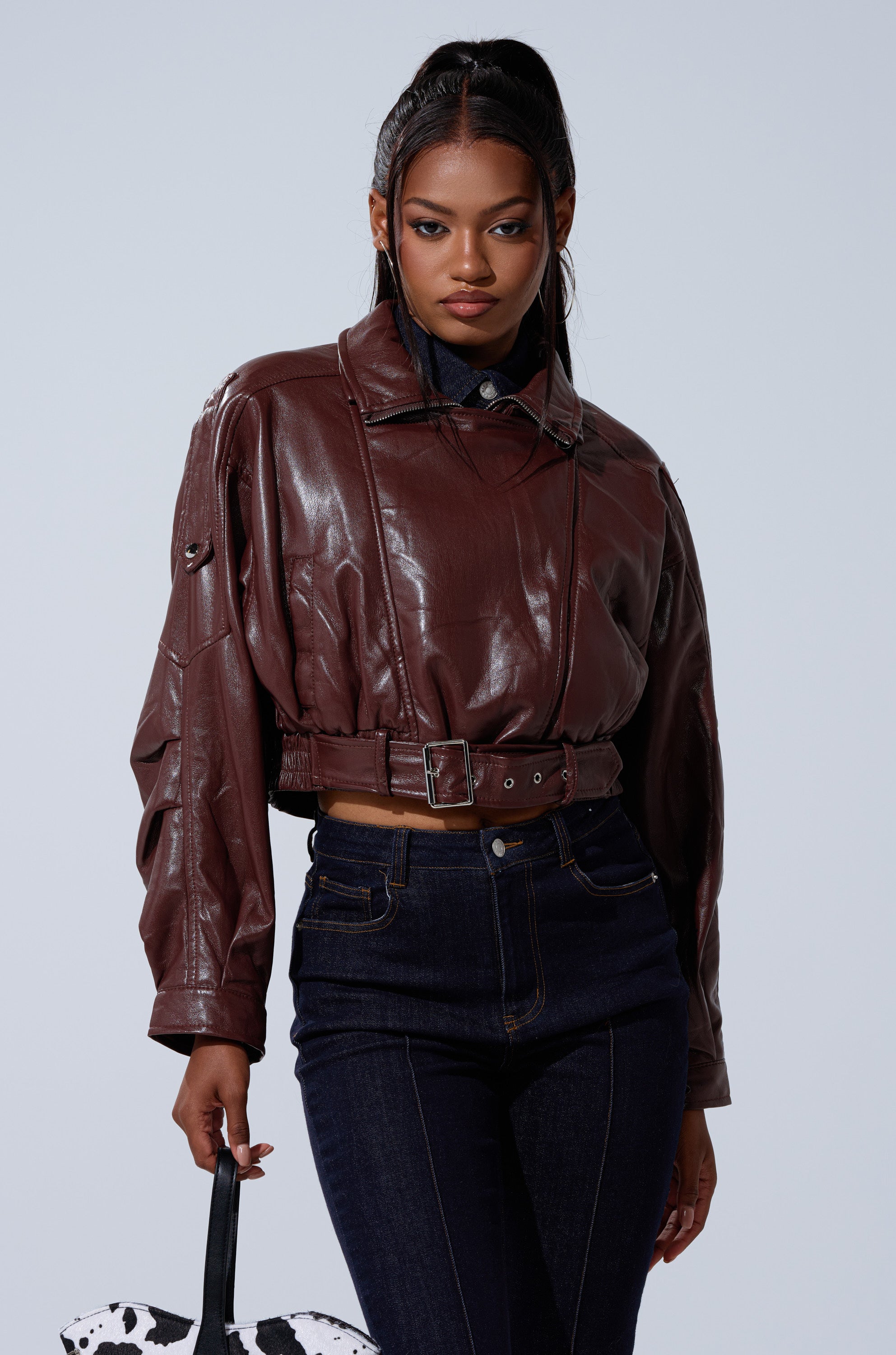 A woman with long dark hair wears the shiny brown EVA SLOUCHY MOTO jacket with dark high-waisted jeans, holding a black and white patterned bag against a plain light background. 4