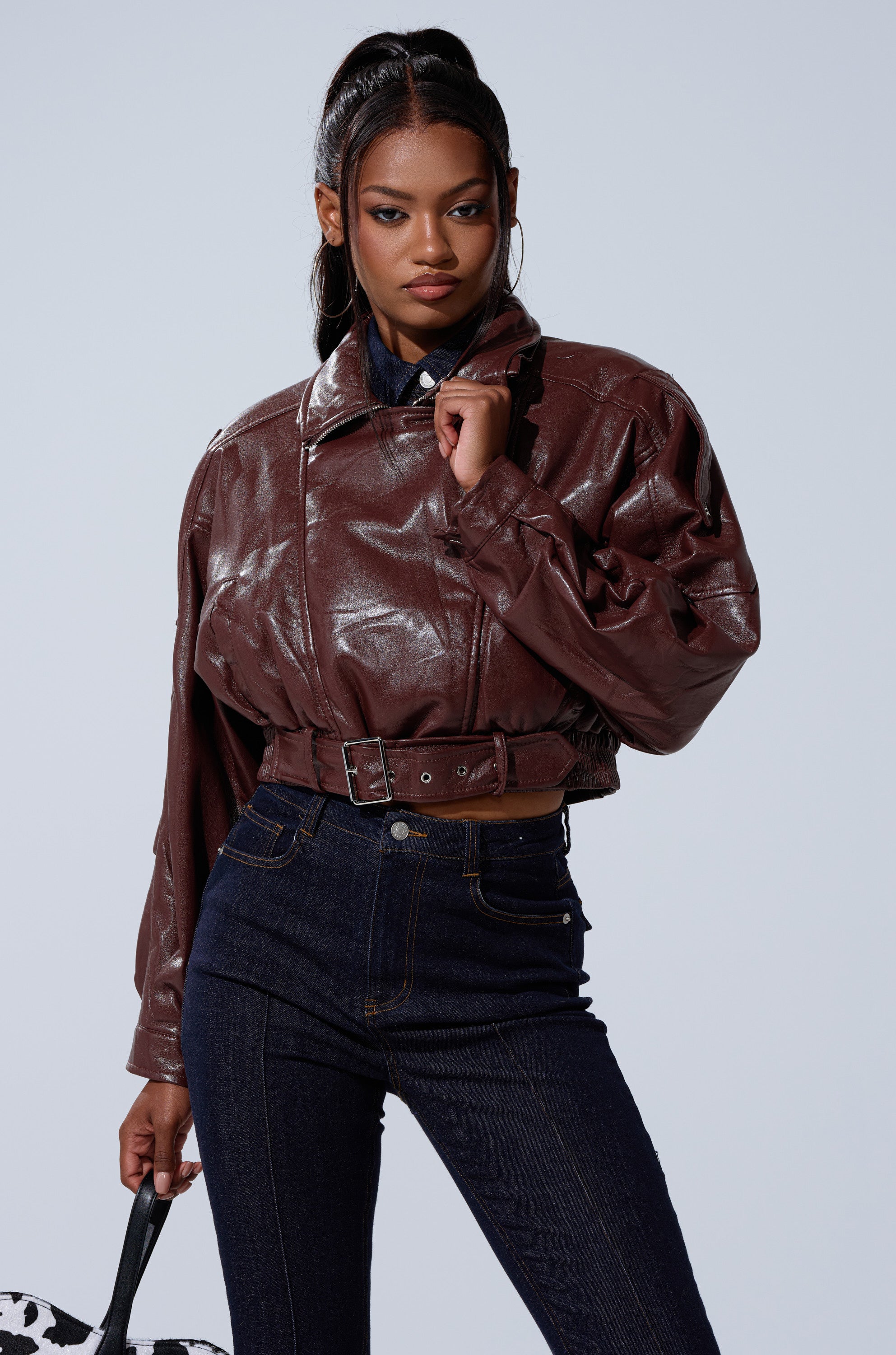 A woman stands confidently against a plain light background, wearing the EVA SLOUCHY MOTO—a shiny brown faux leather moto jacket with a belt—paired with high-waisted dark jeans and holding a black-and-white patterned bag. 0