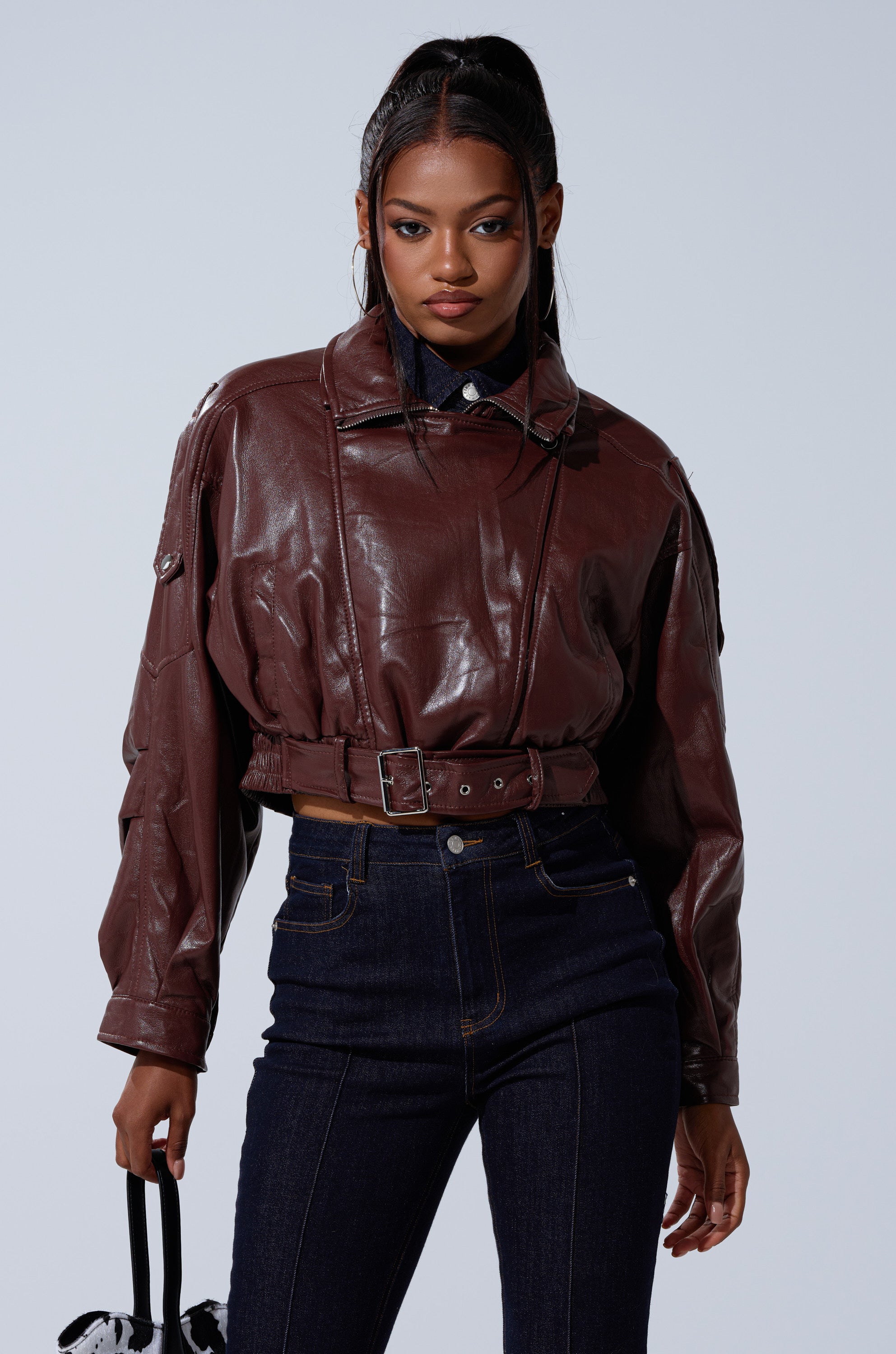 A woman with a high ponytail wears the EVA SLOUCHY MOTO faux leather jacket with a brown belt and high-waisted dark jeans, holding a small black and white bag, standing against a plain light background. 5