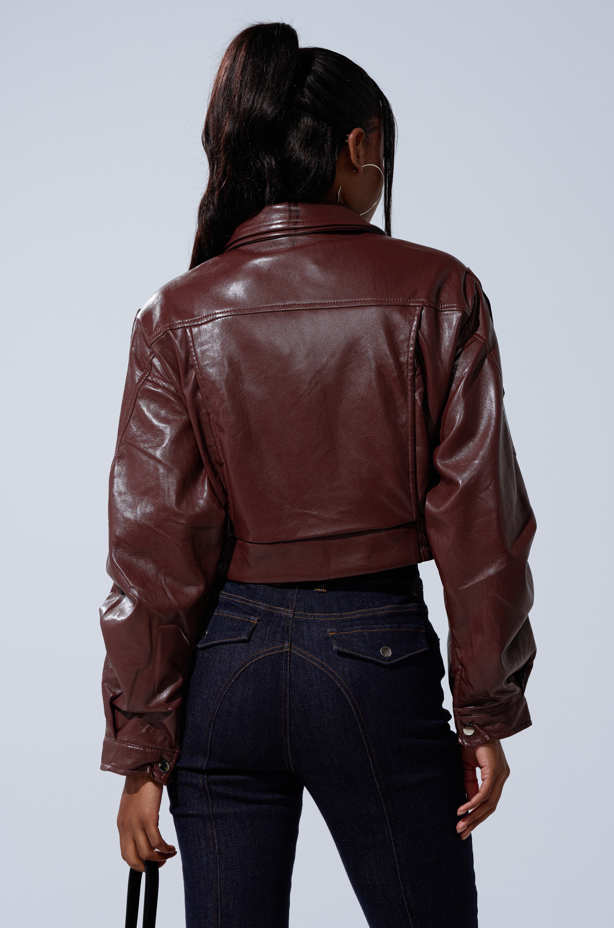 A woman faces away in the EVA SLOUCHY MOTO, a shiny brown faux leather jacket, paired with dark blue jeans. Her long hair is in a high ponytail, and she holds a black bag against a plain light background. 7