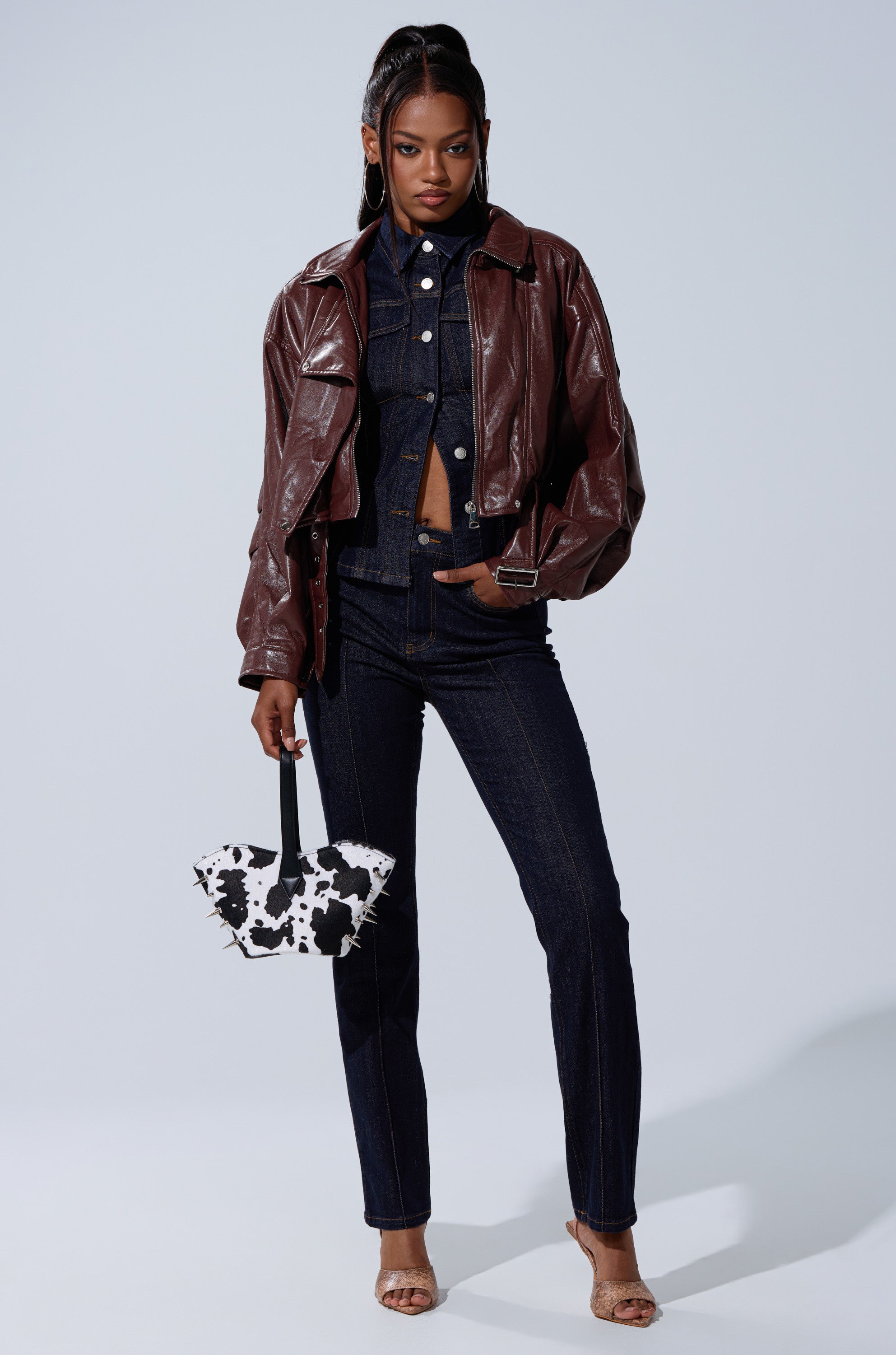 A woman stands confidently in the EVA SLOUCHY MOTO jacket over dark denim, holding a black and white cow print purse and wearing open-toe heels against a light background. 8