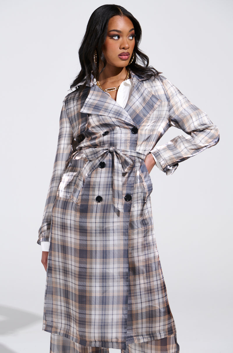 A confident woman stands with one hand on her hip, wearing the "PLAID ABOUT YOU SHEER TRENCH" coat adorned with large buttons and a tied belt. The longline silhouette complements her white shirt, flowing wavy hair, and gold hoop earrings against a plain light gray background.