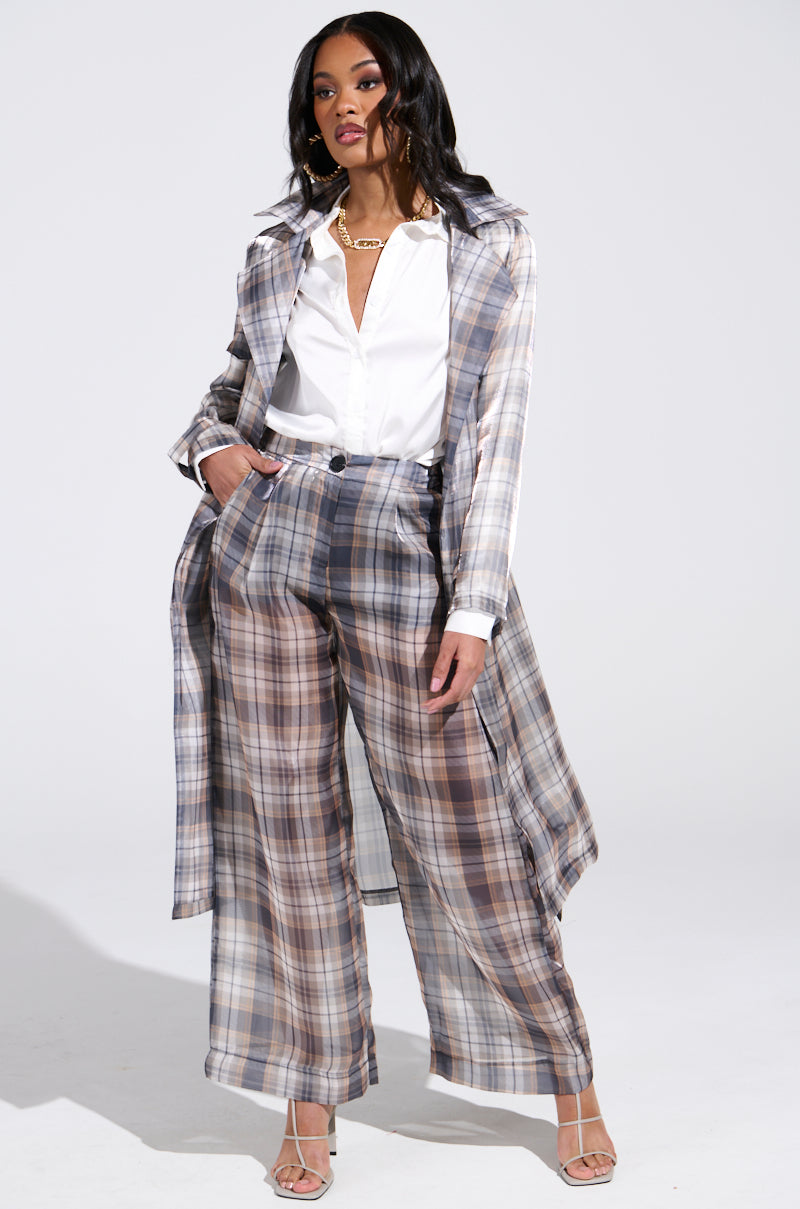 A woman confidently poses in the PLAID ABOUT YOU SHEER TRENCH over a white blouse and wide-leg pants. She pairs this ensemble with nude heels and a gold necklace, standing against a neutral backdrop to showcase the longline silhouette of her stylish choice.