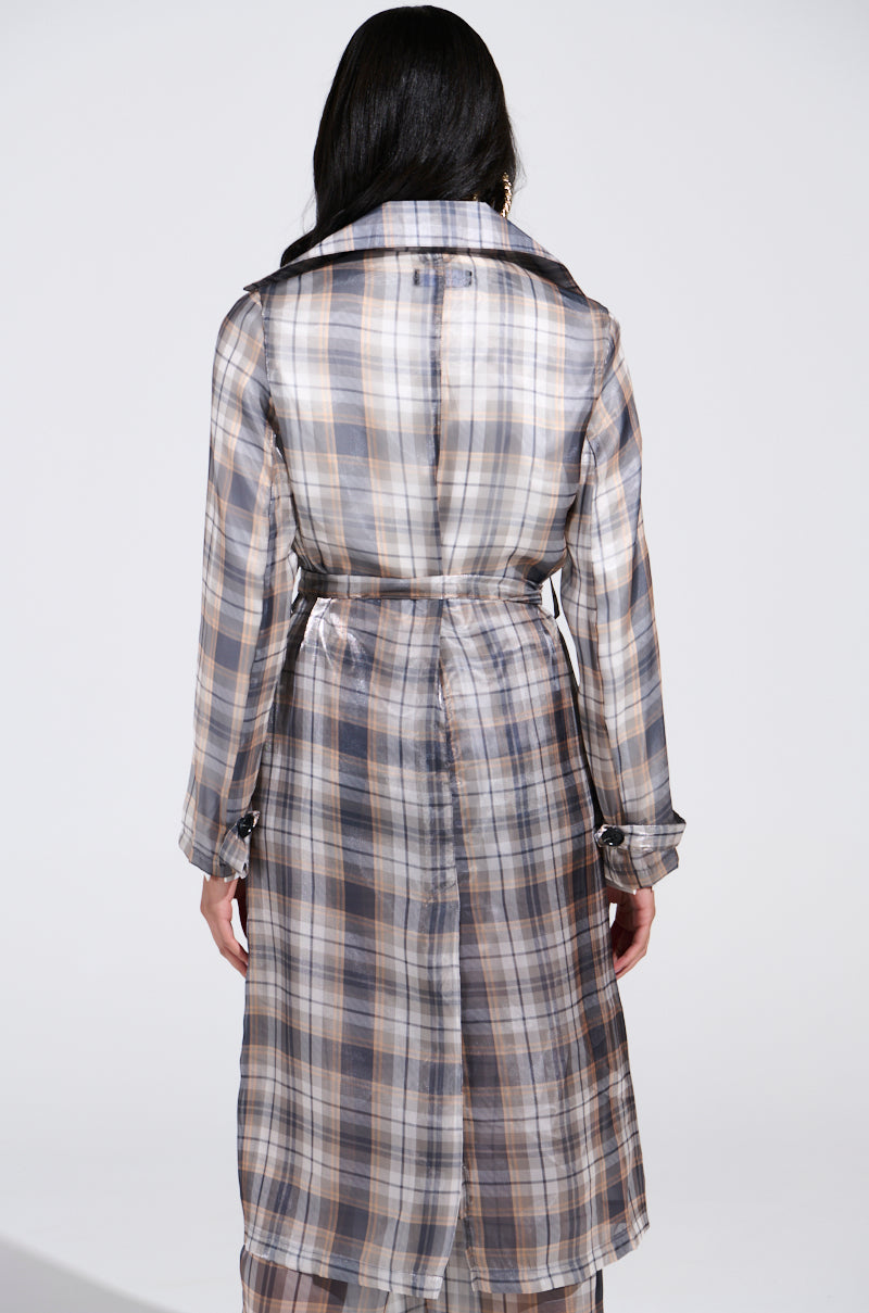 A person with long dark hair, facing away, dons the "Plaid About You Sheer Trench," which showcases a longline silhouette in blue, brown, and white plaid. It's belted at the waist and set against a plain white backdrop.