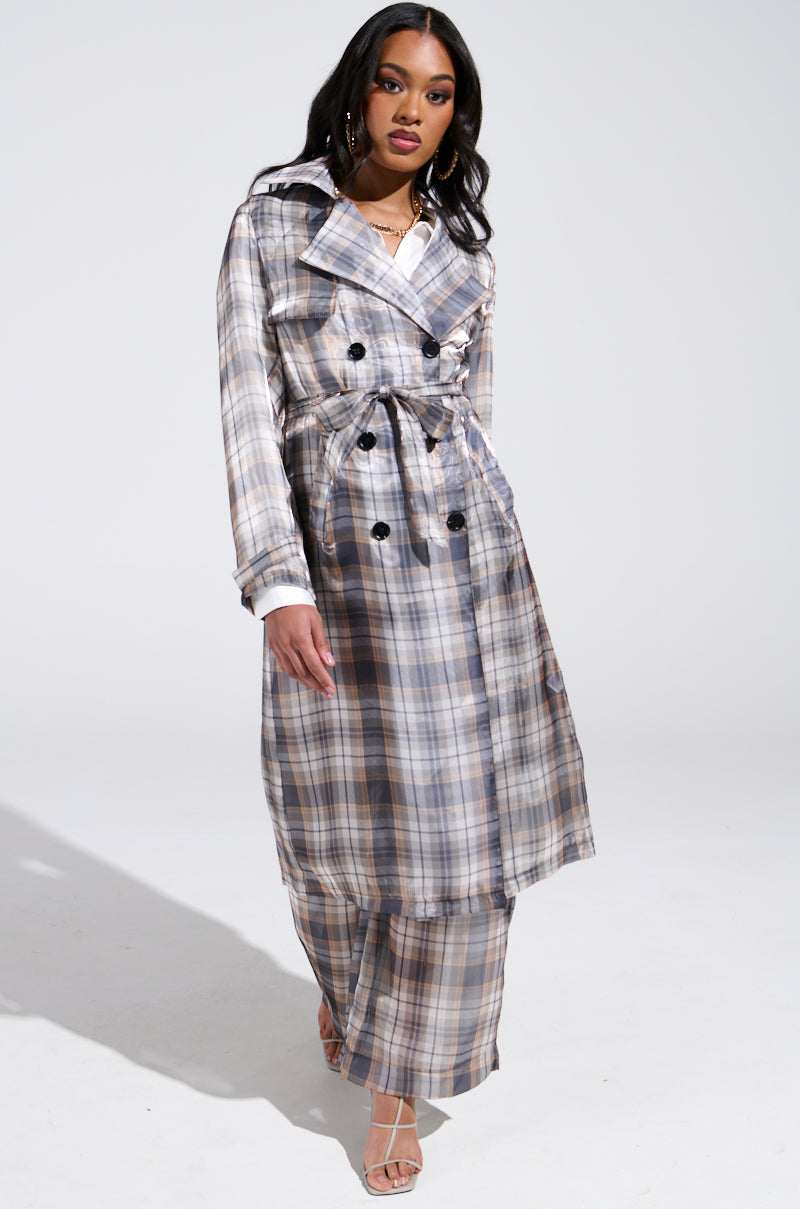 A woman models the PLAID ABOUT YOU SHEER TRENCH, a longline plaid coat with double-breasted design, belted waist, and full-length sleeves. She pairs it with matching wide-leg trousers and strappy heels on a plain white background.