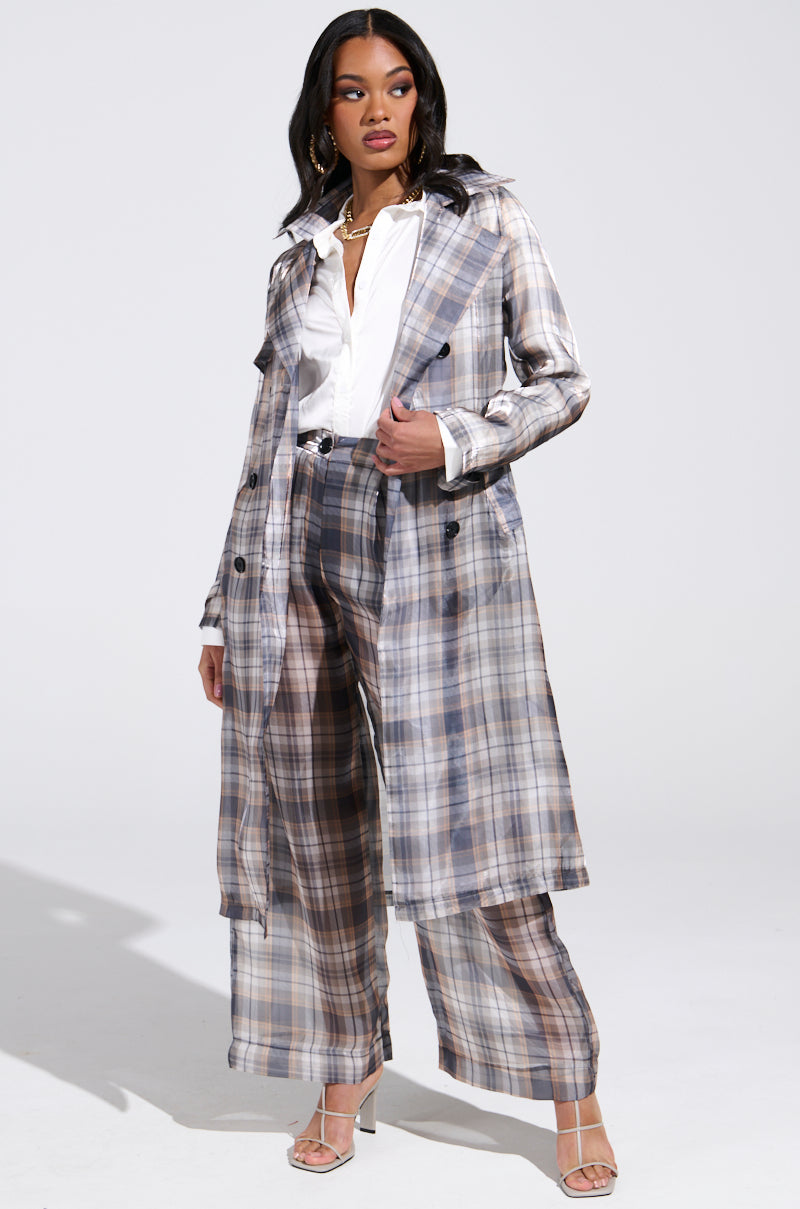 A person confidently wears the "Plaid About You Sheer Trench," a stylish plaid coat with matching wide-leg pants. They pair it with a white shirt, a necklace, and open-toed heels against a plain background.