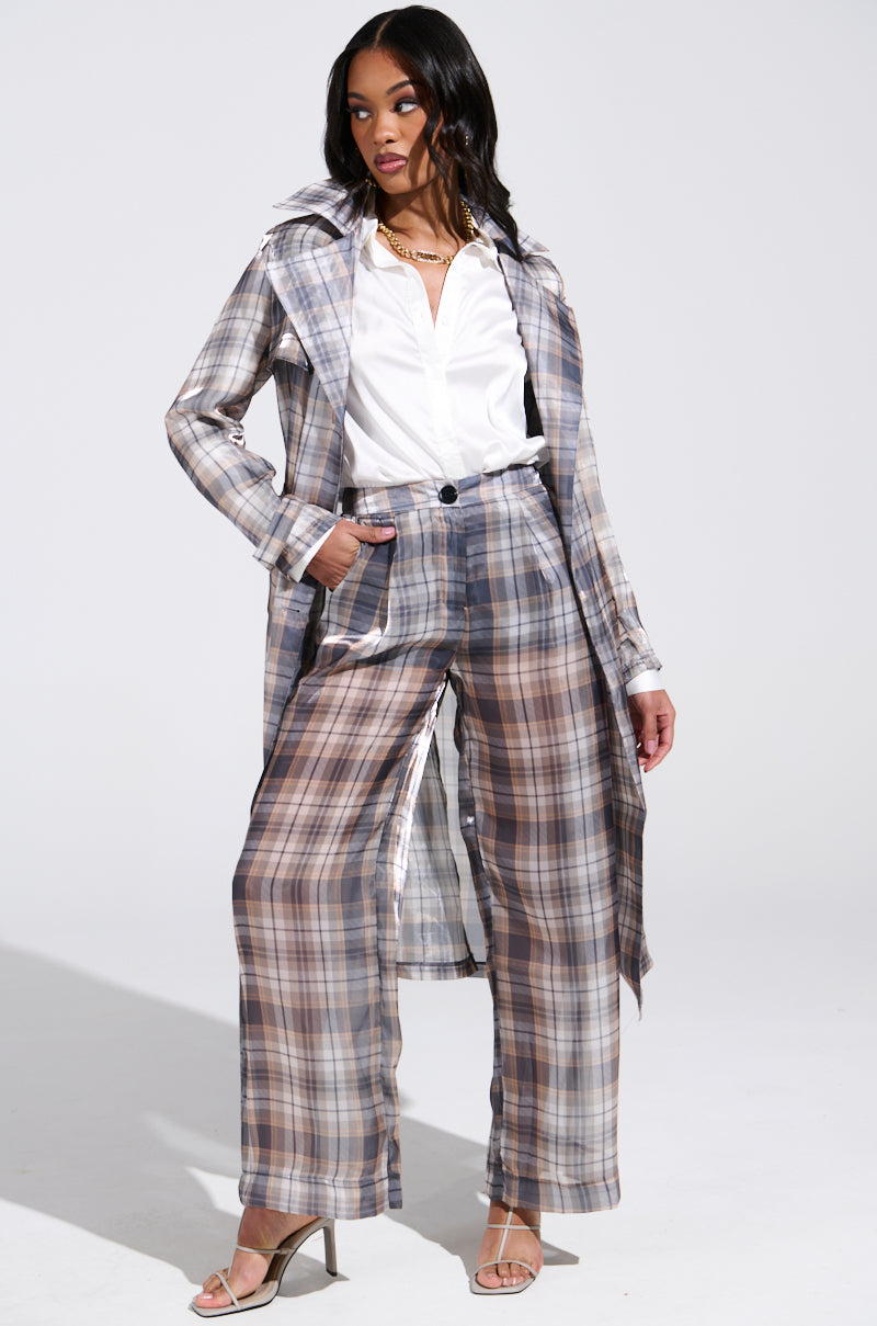 A person models the "PLAID ABOUT YOU SHEER TRENCH" featuring a longline silhouette. It's stylishly paired with matching wide-legged pants, a crisp white shirt, and high-heeled sandals; one hand rests in their pocket against a plain white background.