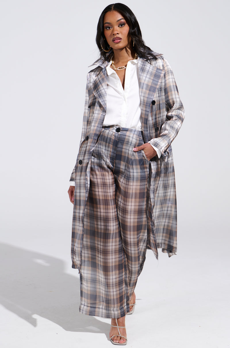 Wearing the "Plaid About You Sheer Trench" with wide-legged plaid pants and a white shirt, they confidently strut with long dark hair perfectly complementing a gold chain necklace and open-toe heels against a plain white backdrop.