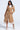 Wearing the SOMETHING SWEET BUTTER LUXE TRENCH over a white shirt and light skirt, a person stands with one hand in their pocket. Their look is completed by strappy high-heeled shoes against a plain backdrop.
