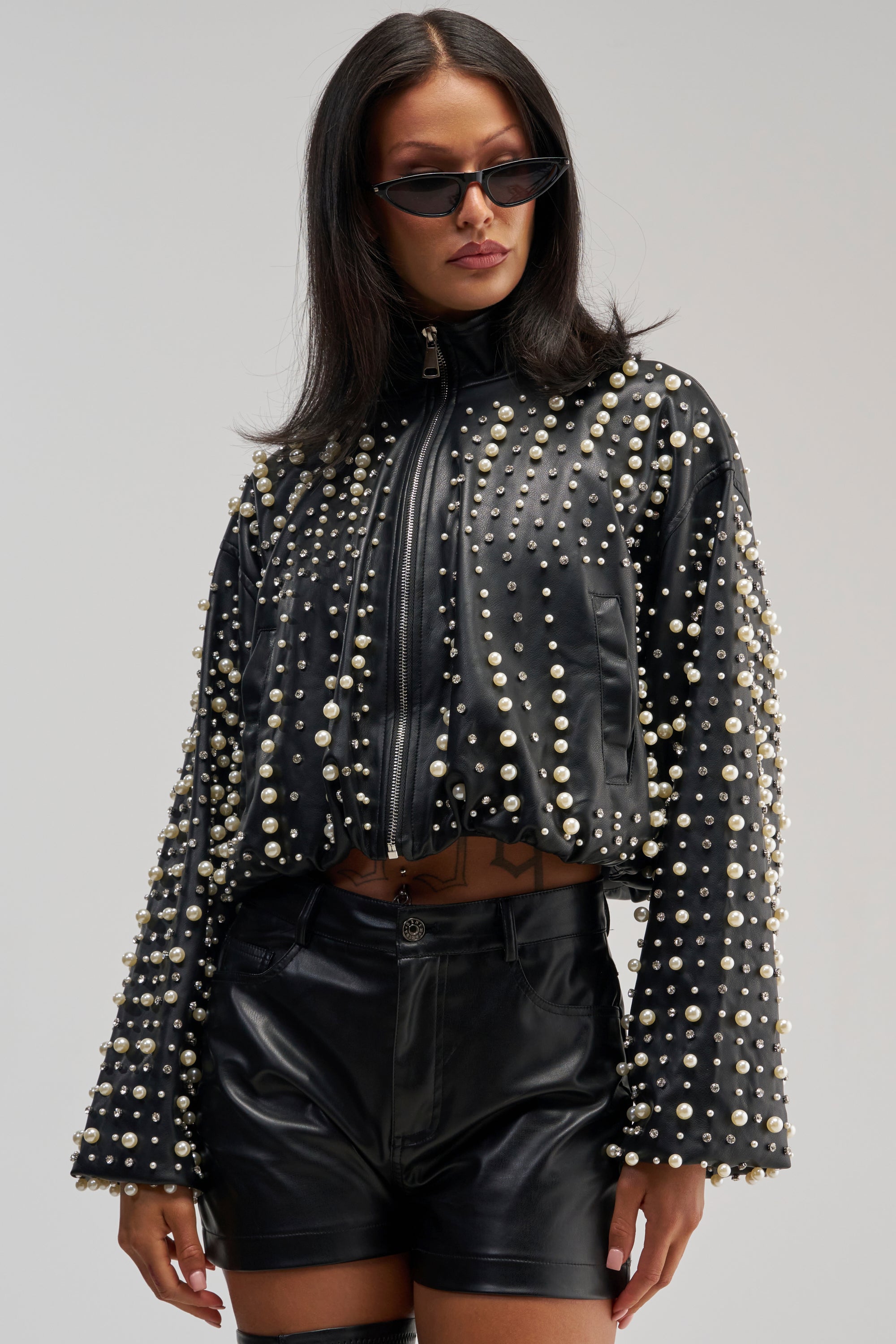 Wearing the EUPHORIA EMBELLISHED BOMBER, black leather shorts, and cat-eye sunglasses, a woman stands confidently against a plain background—creating a striking streetwear look.