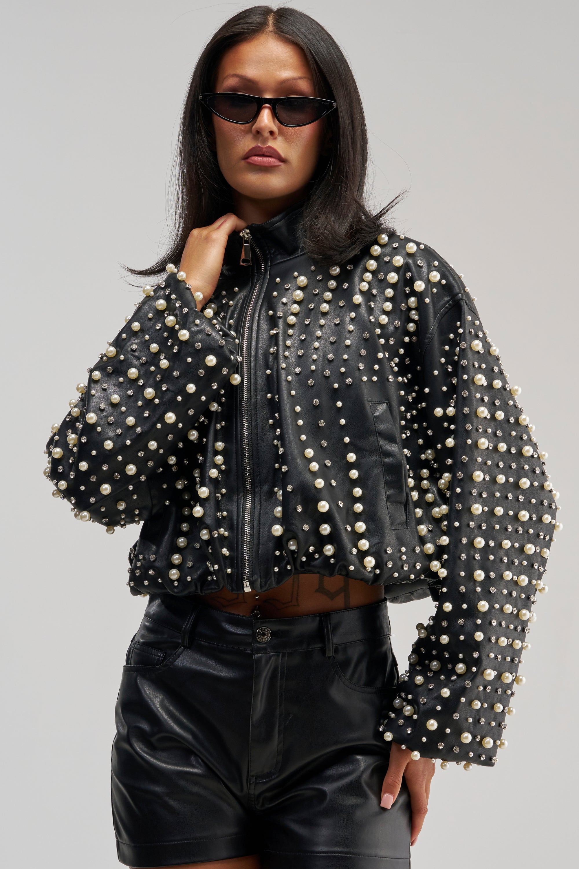 Dressed in the EUPHORIA EMBELLISHED BOMBER adorned with pearl details and paired with black leather shorts and dark sunglasses, the woman poses confidently against a plain background, embodying bold streetwear style.