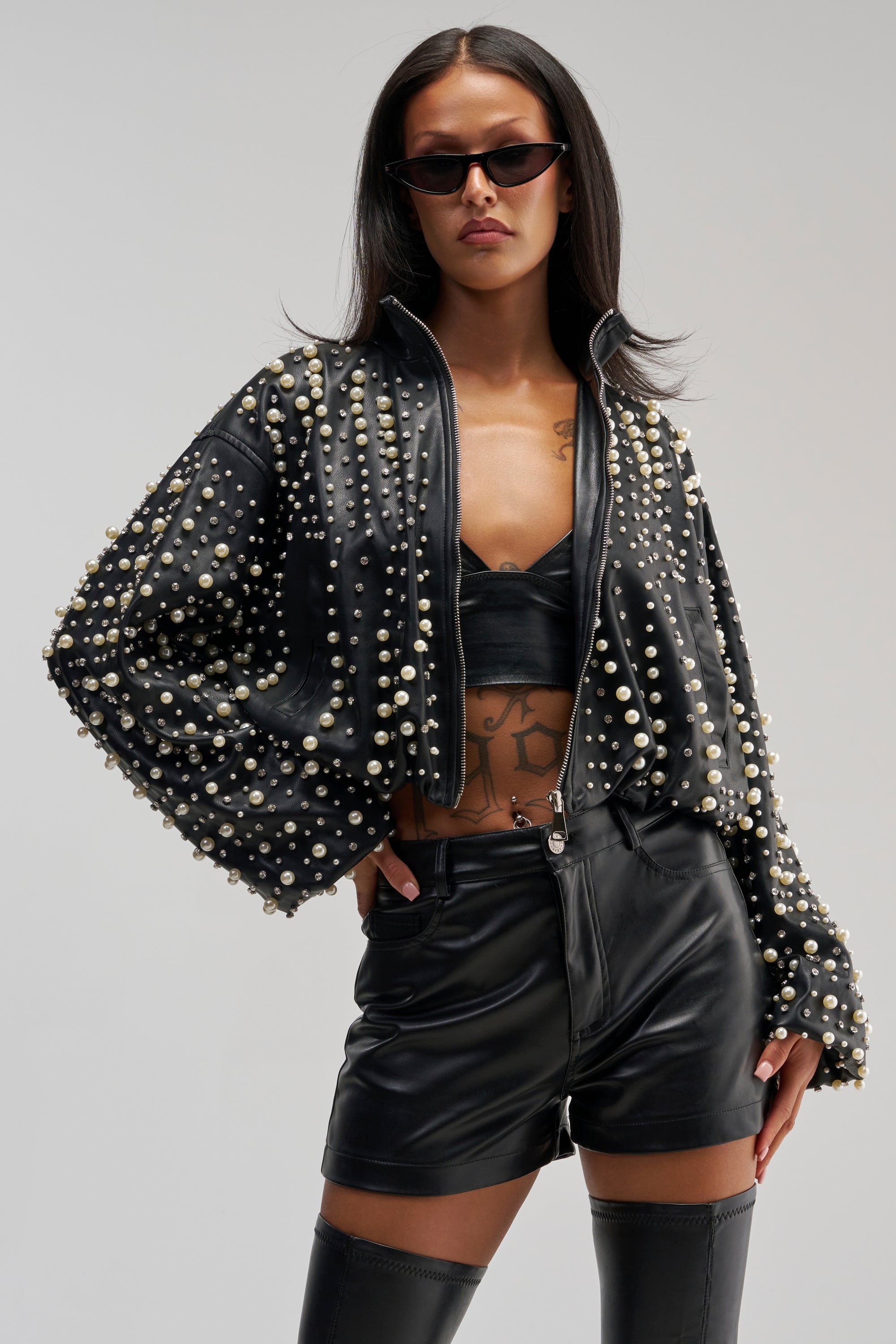 A woman wearing black sunglasses, the EUPHORIA EMBELLISHED BOMBER styled in a pearl-studded black leather design, a black bralette, black leather shorts, and thigh-high boots poses confidently with one hand on her hip against a plain background.