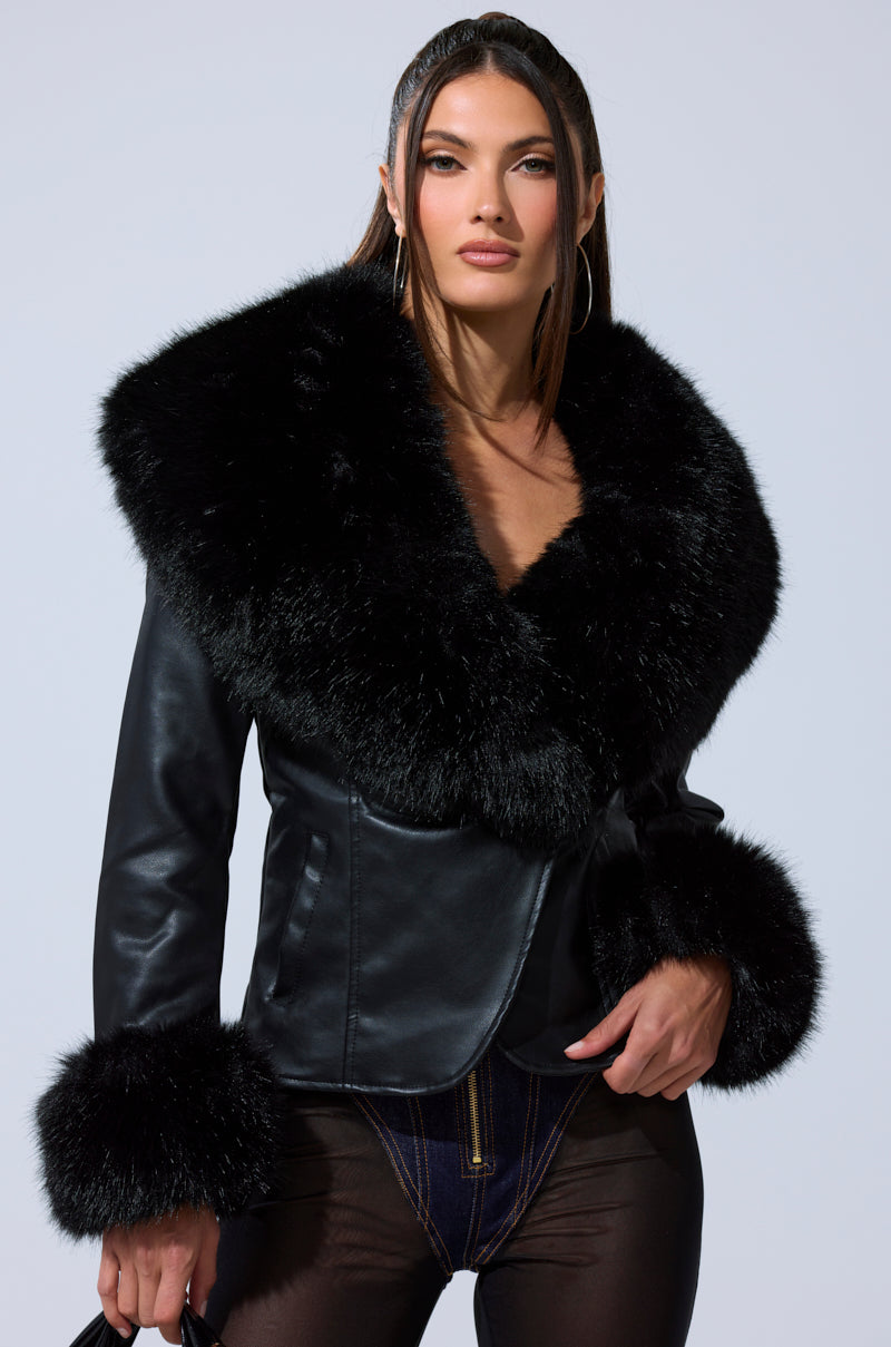 A woman with long, straight brown hair poses confidently against a light background, wearing the CELESTE FUR TRIM BLAZER with plush black fur accents on the collar and cuffs, and holding a small black handbag.