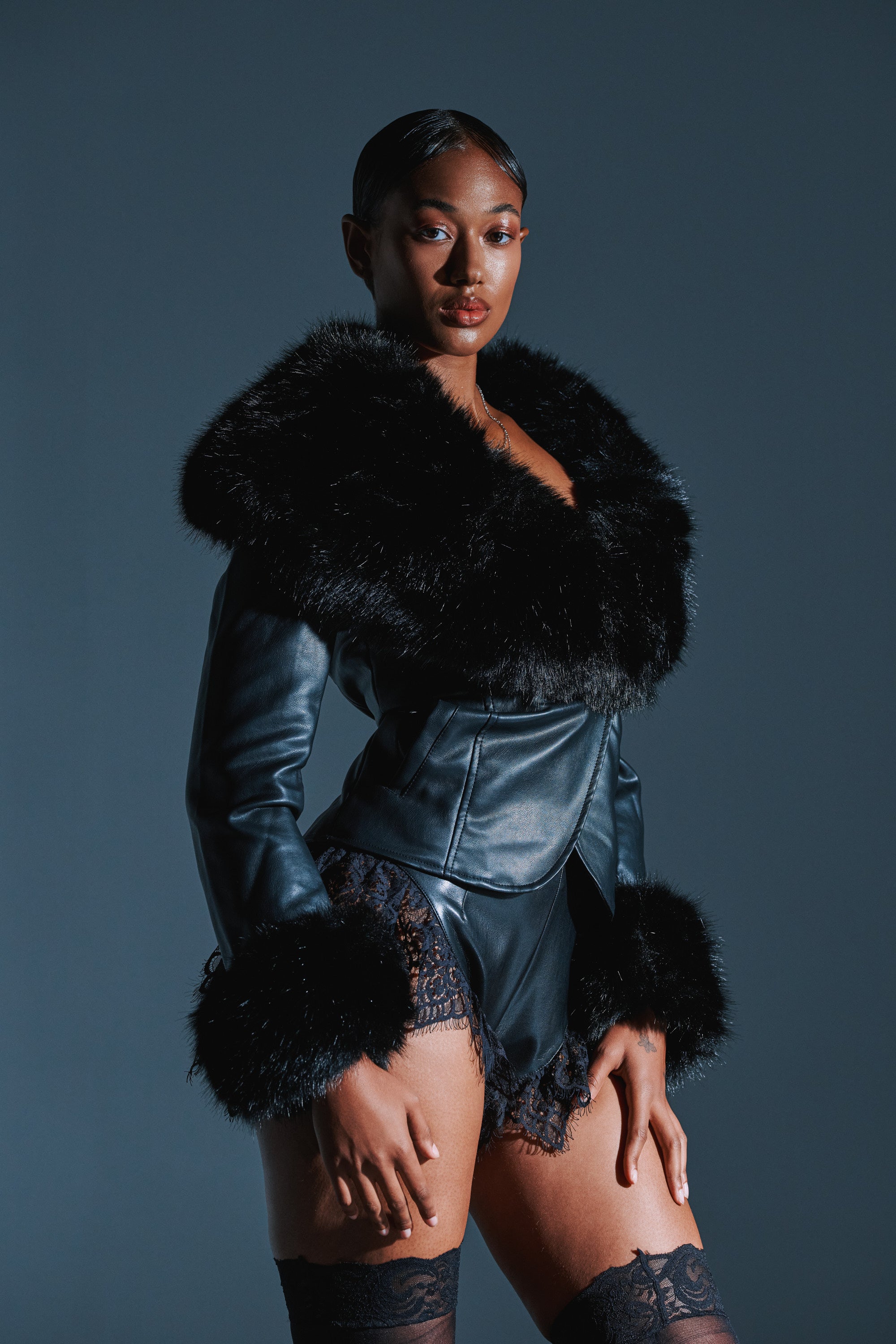 A woman poses confidently in the CELESTE FUR TRIM BLAZER, featuring dramatic faux fur, paired with black lace shorts and thigh-high stockings. Her sleek, pulled-back hair stands out against the dark background.