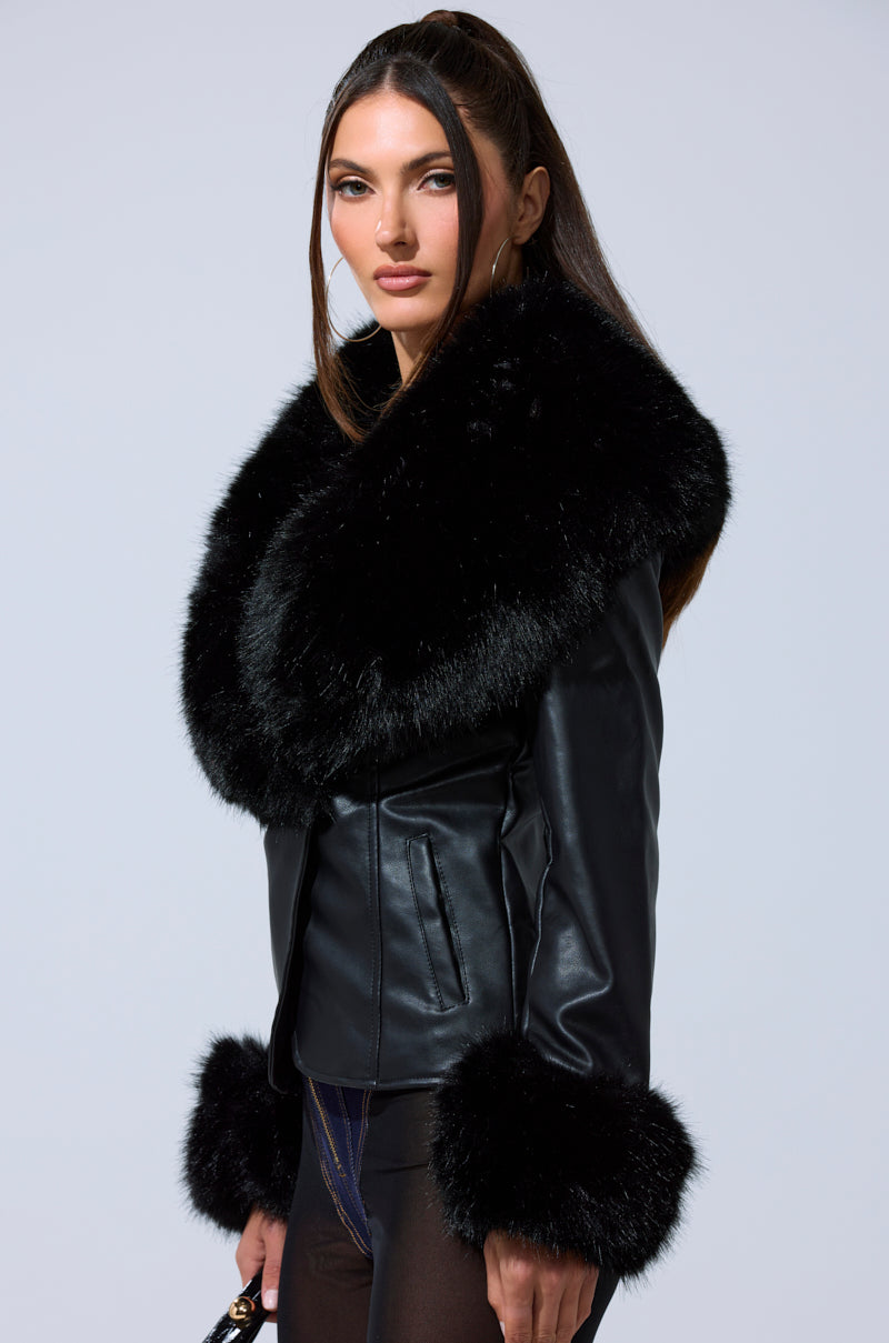 A woman with long brown hair models the CELESTE FUR TRIM BLAZER, featuring large, fluffy black fur on the collar and cuffs, paired with dark pants against a light gray background.