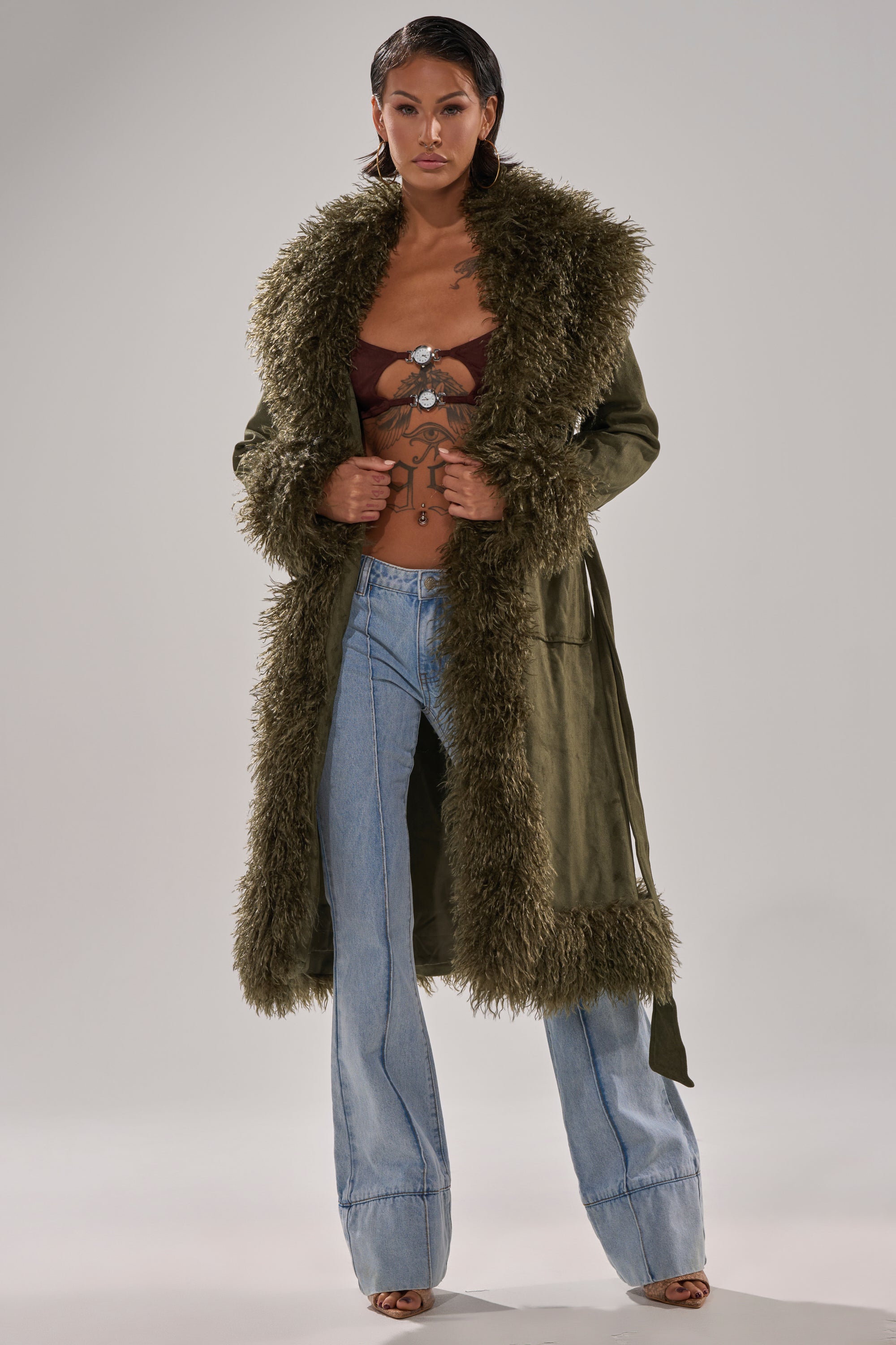 Confidently posing against a neutral background, a person models the JADE FUR TRIM TRENCH—a long faux fur trench coat with shaggy trim—paired with blue flared jeans and a brown bralette, showcasing visible tattoos.