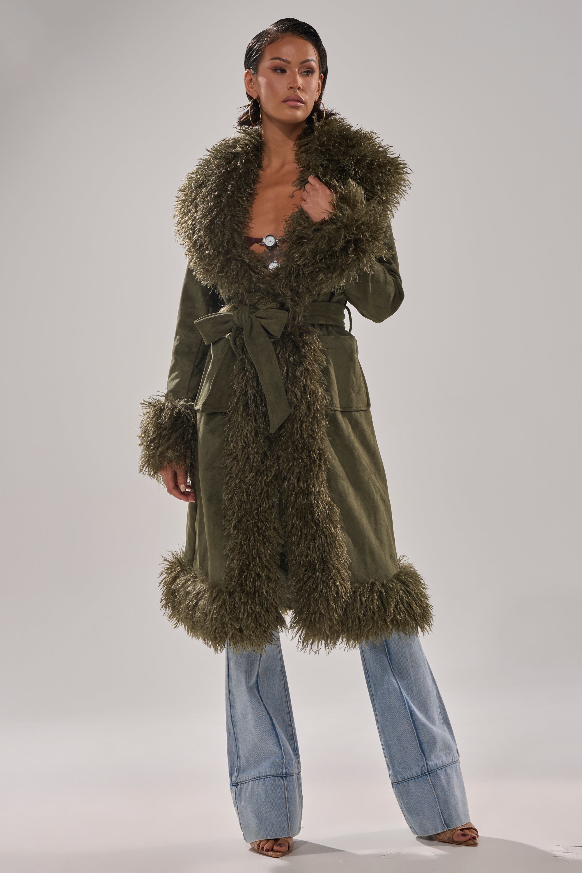 A woman models the JADE FUR TRIM TRENCH, featuring a fluffy faux fur collar, cuffs, and hem. She pairs it with light blue flared jeans and open-toe heels, standing against a plain gray background.