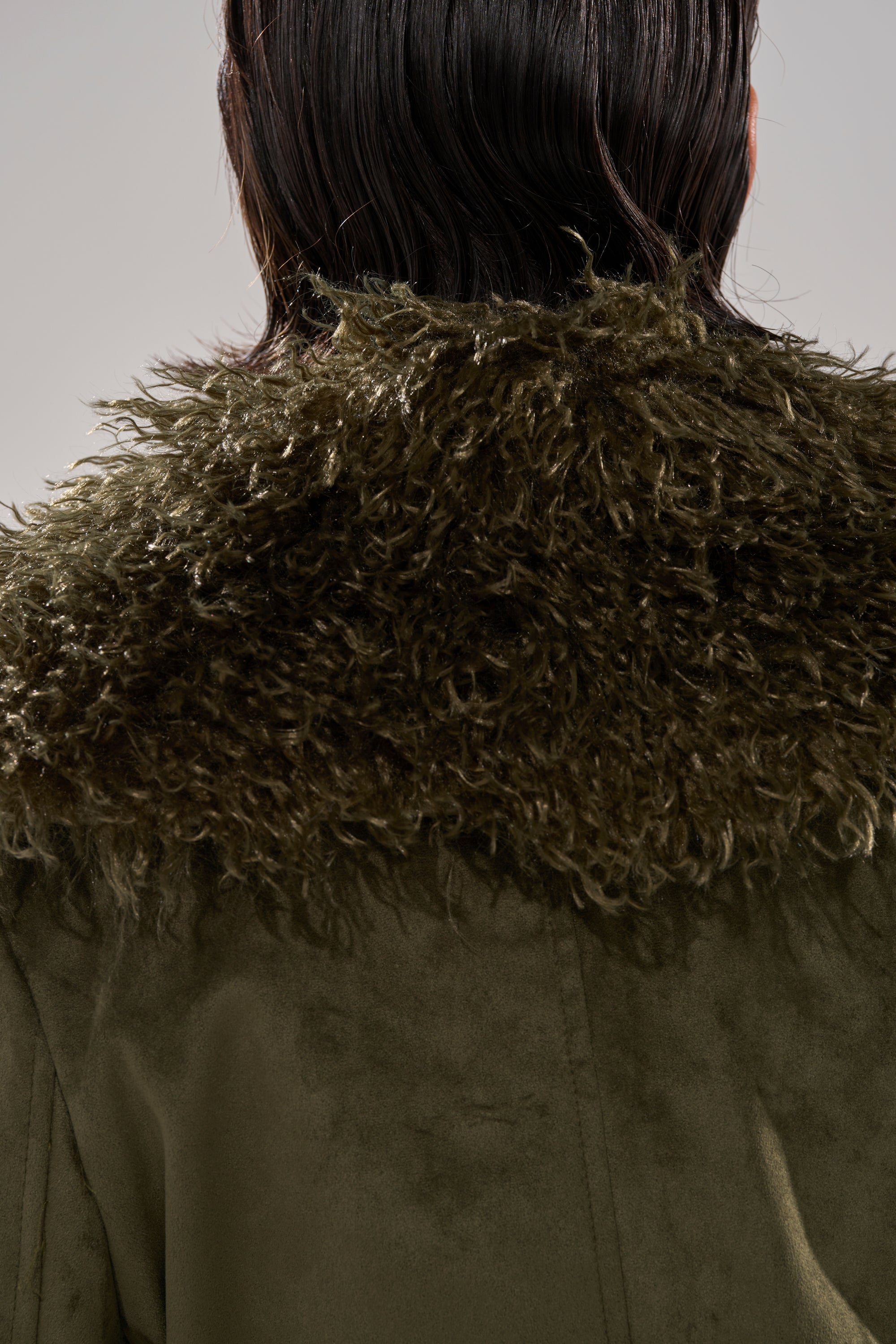 A close-up of a person’s back shows the JADE FUR TRIM TRENCH in olive green, featuring a thick, textured collar with wet, dark hair falling just above it.