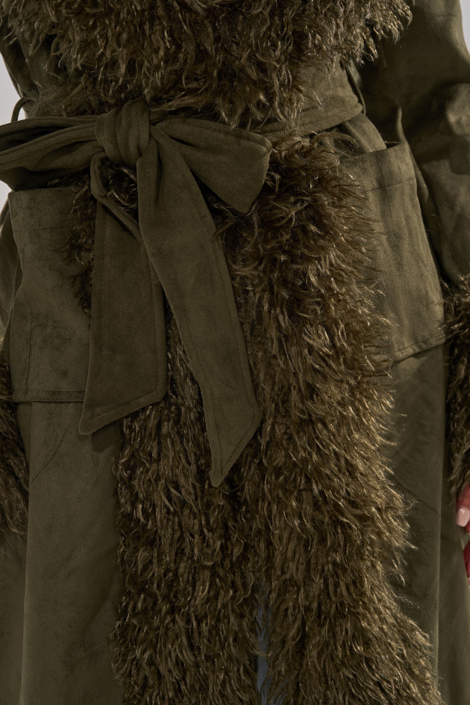 A close-up of a person wearing the JADE FUR TRIM TRENCH, an olive green longline trench coat with a matching fabric belt at the waist and furry textured trim along the front.