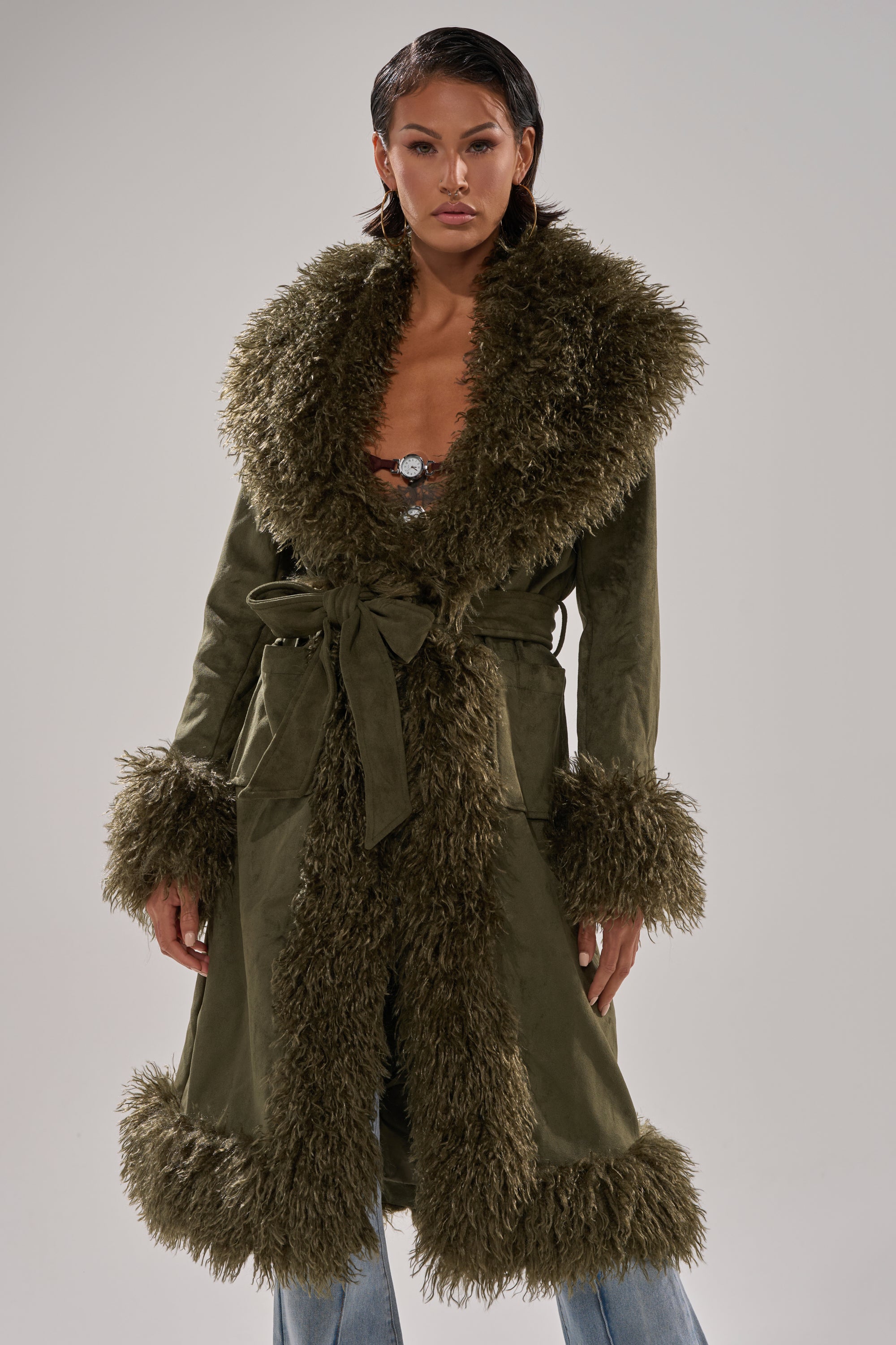 A woman models the JADE FUR TRIM TRENCH in olive green, featuring thick faux fur trim on the collar, cuffs, and hem, belted at the waist and styled over blue jeans against a plain background.