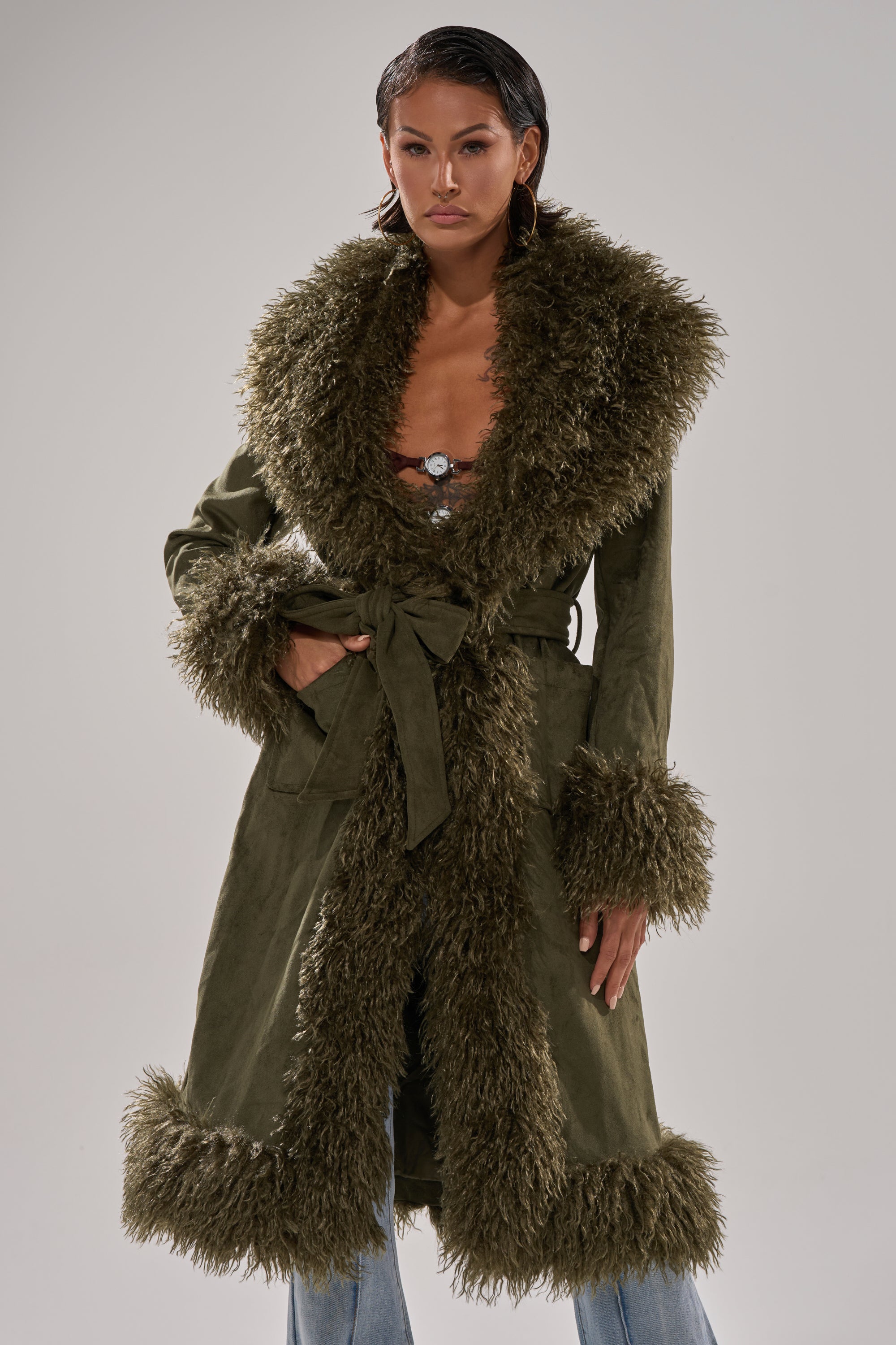 A person wears the JADE FUR TRIM TRENCH in olive green faux suede with large, fluffy fur trim on the collar, cuffs, and hem. The longline coat is partially tied at the waist over blue jeans against a neutral gray background.