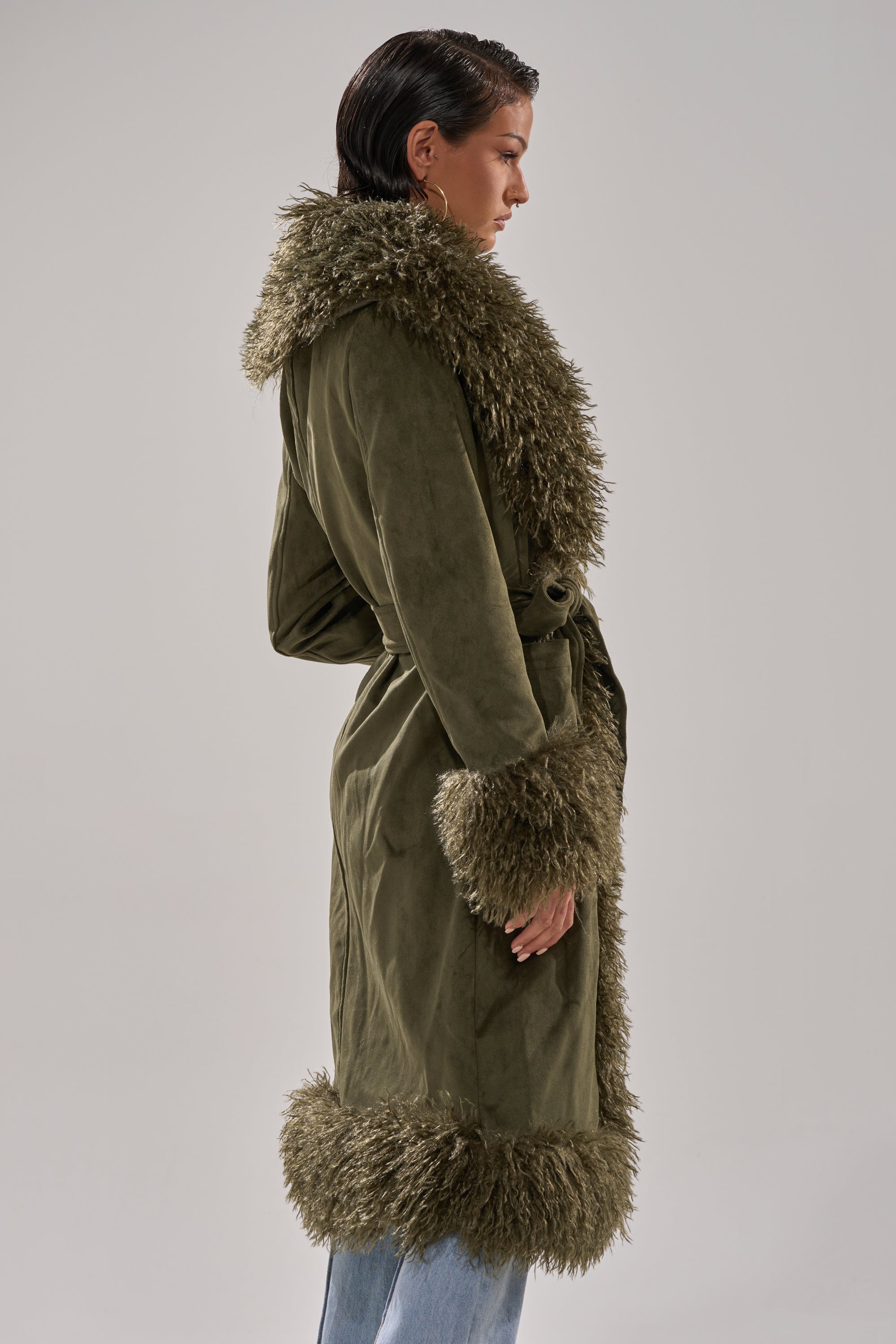 A person in profile models the JADE FUR TRIM TRENCH, a faux suede coat with fur collar, cuffs, and hem. The long, belted silhouette is styled with light blue jeans and short, slicked-back hair for a chic look.