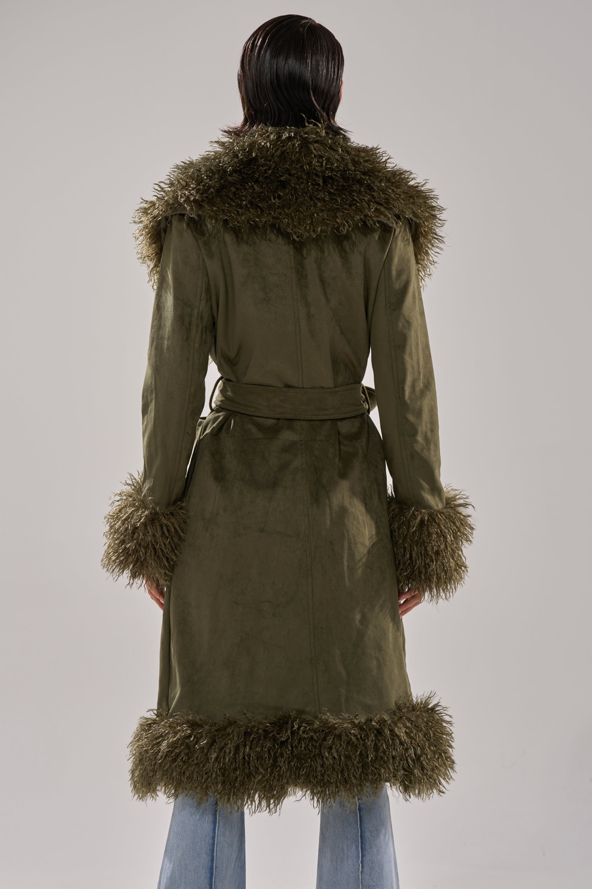 Seen from the back, a person wears the JADE FUR TRIM TRENCH in olive-green, featuring fur trim on the collar, cuffs, and hem, a belted waist, longline silhouette, and is styled with light blue jeans.