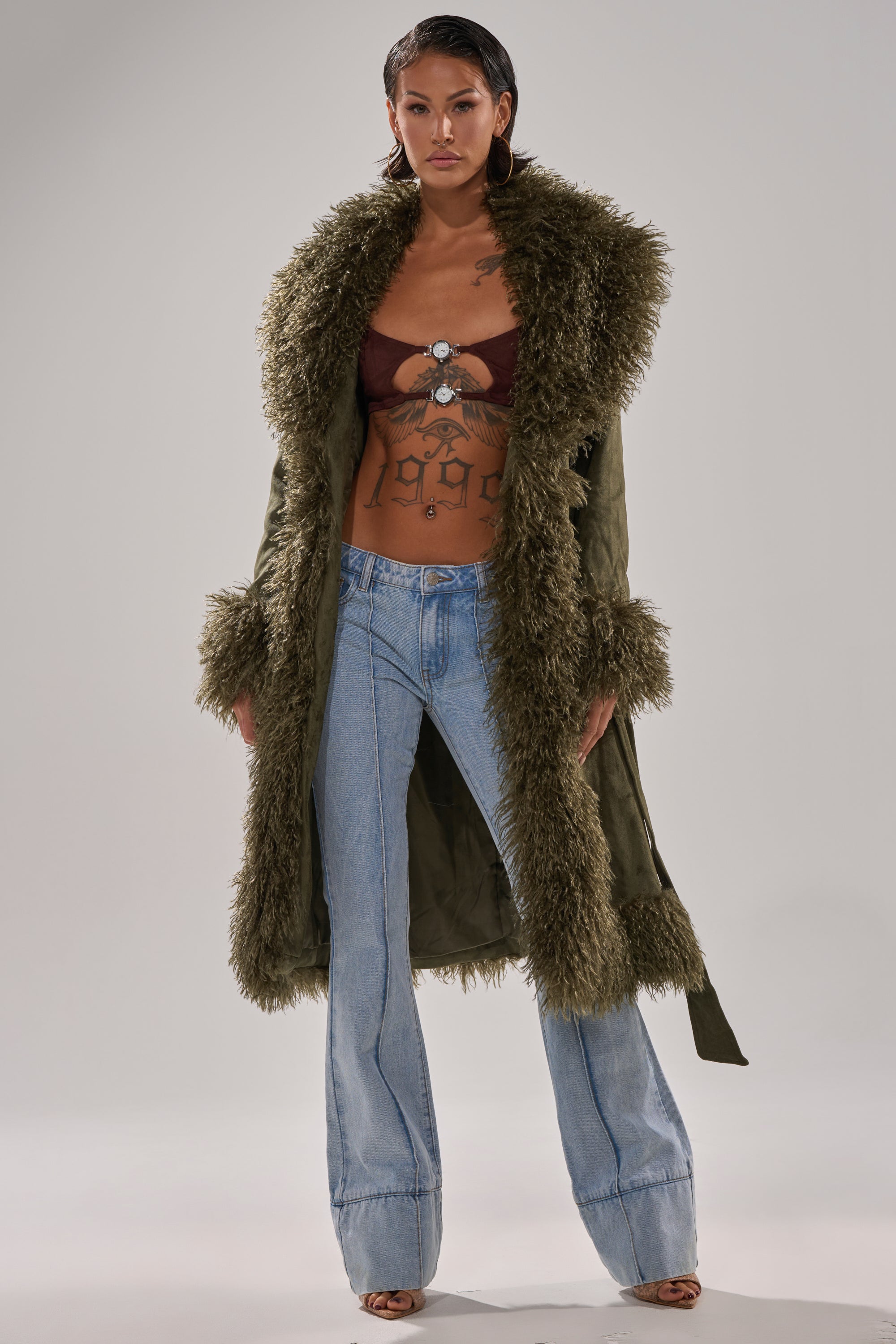 A person stands confidently in light blue flared jeans, a maroon bralette, and the olive JADE FUR TRIM TRENCH by AKIRA Label with shaggy fur at the collar, cuffs, and hem. They have tattoos and wet-look hair.
