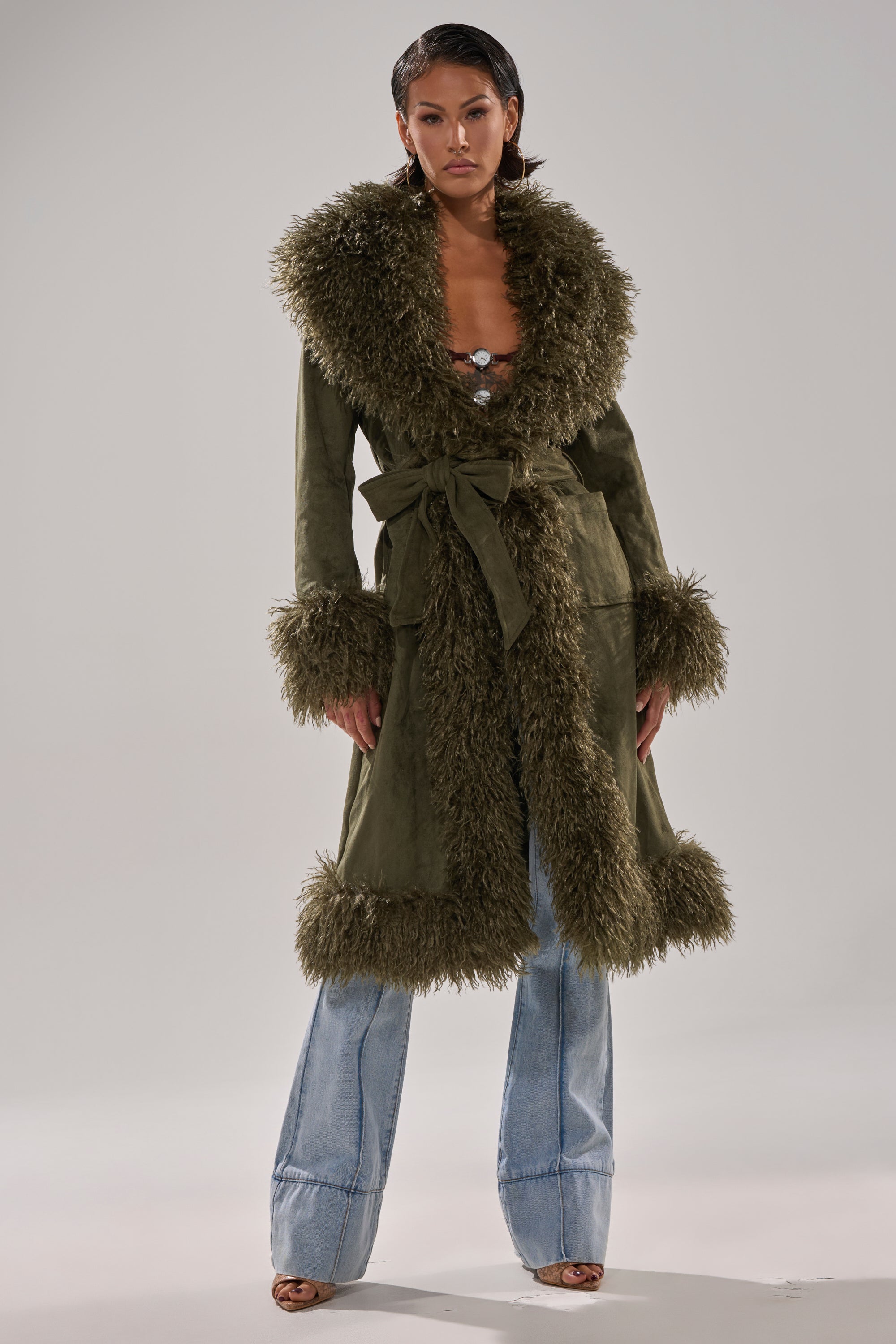 A woman wears the JADE FUR TRIM TRENCH, a longline olive green faux suede coat with shaggy fur trim on the collar, cuffs, and hem, belted at the waist over a patterned top and flared light blue jeans.