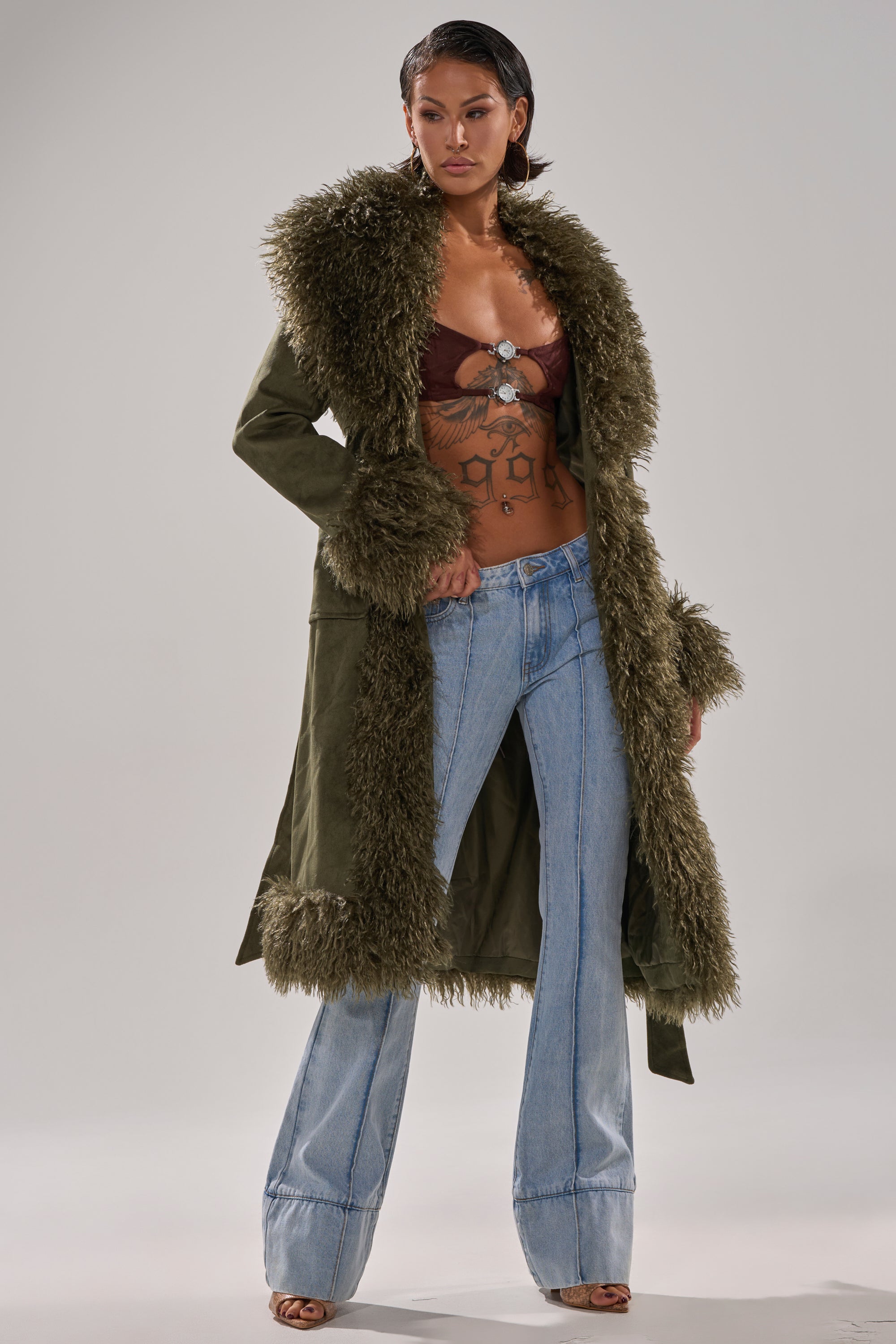 Wearing the JADE FUR TRIM TRENCH in olive green, paired with a brown bra top, light blue flared jeans, and open-toed heels, a person stands confidently against a plain background.