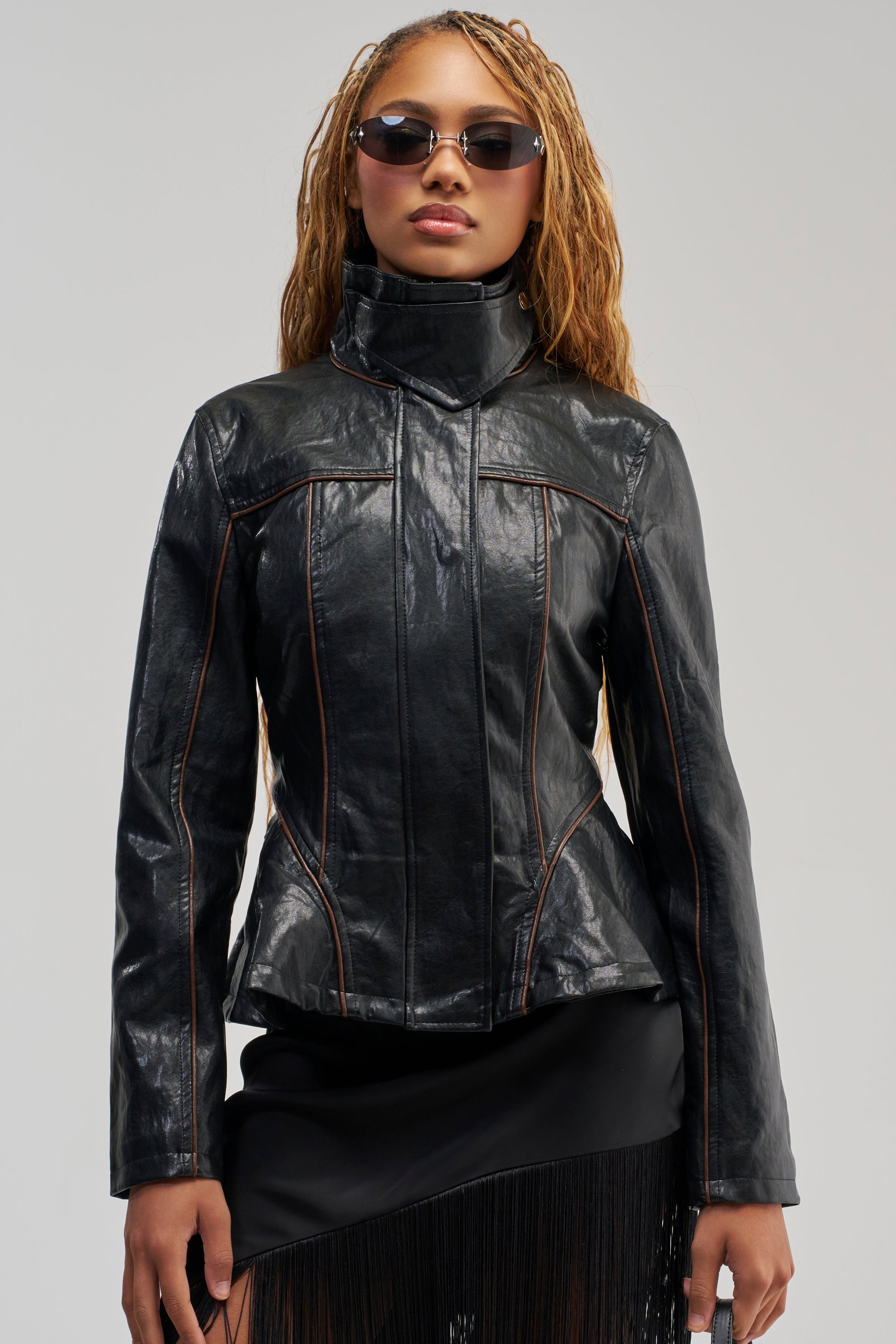 A person wearing sunglasses, the SMALL CIRCLE PEPLUM MOTO—a high-collared black moto jacket with brown seams—and a black skirt with fringe stands facing forward against a plain background.