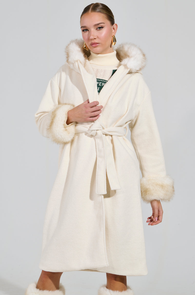 A person wearing the LILA FUR TRIM COAT, featuring a long design with a hood and faux fur cuffs, stands confidently against a plain background. The coat is elegantly tied with a belt around the waist.