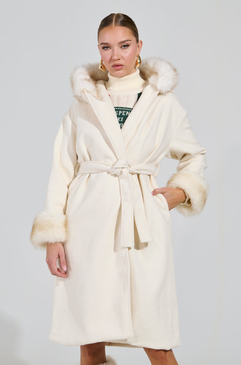 A person wearing the sophisticated LILA FUR TRIM COAT, featuring a cream-colored design with a fur-lined hood and cuffs. The coat is elegantly belted at the waist, and they exude confidence while standing against a plain background.