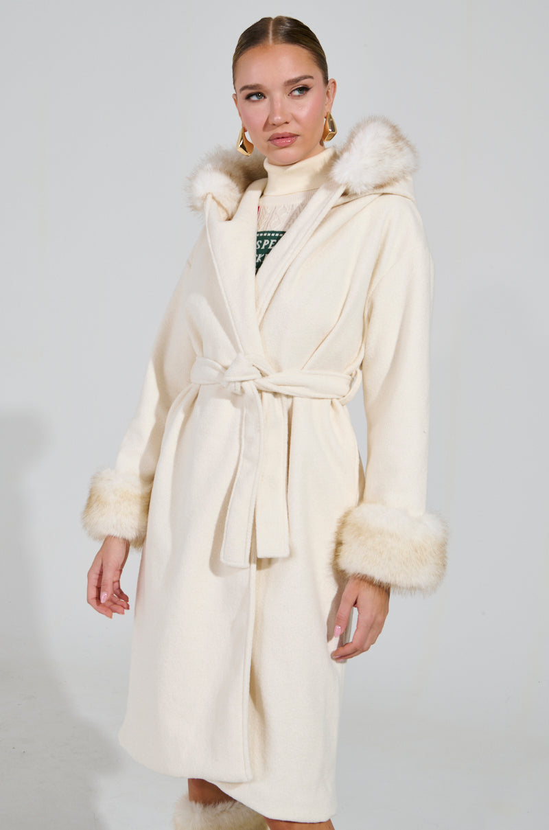 A woman is dressed in the LILA FUR TRIM COAT, which features a cream color with a belt and faux fur trim on the hood and cuffs. Her hair is tied back, and she complements the outfit with a turtleneck sweater underneath. The plain, light-colored background enhances the elegance of her attire.