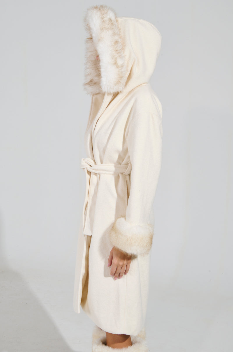 A person stands sideways in the elegant LILA FUR TRIM COAT, showcasing its cream color, belt, and stylish faux fur cuffs against a plain white backdrop.