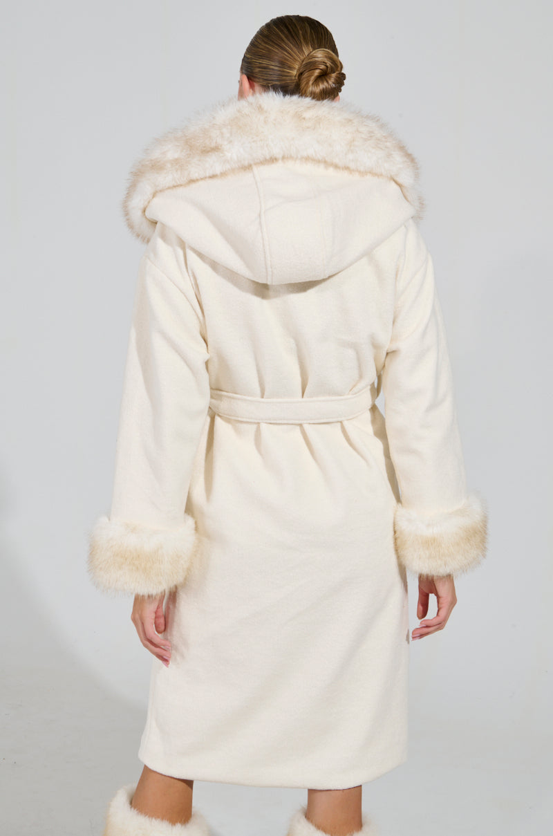 A person is wearing the LILA FUR TRIM COAT, featuring a cream color with a hood and faux fur accents on the cuffs and hem. The belted waist adds definition to the ensemble as the individual, facing away, highlights a tidy bun hairstyle.